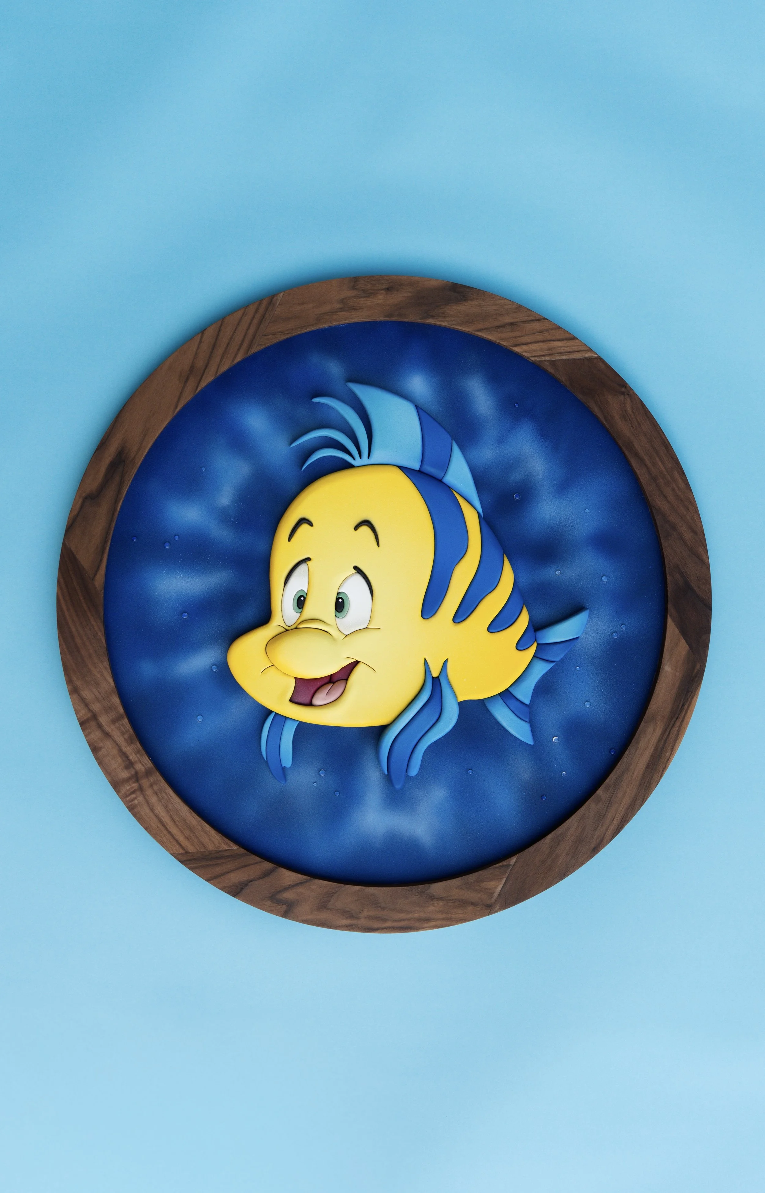 Decorative wall art of a cartoon yellow fish with blue stripes and fins, smiling against a blue background, framed with a wooden circle. Flounder wood art. The little mermaid