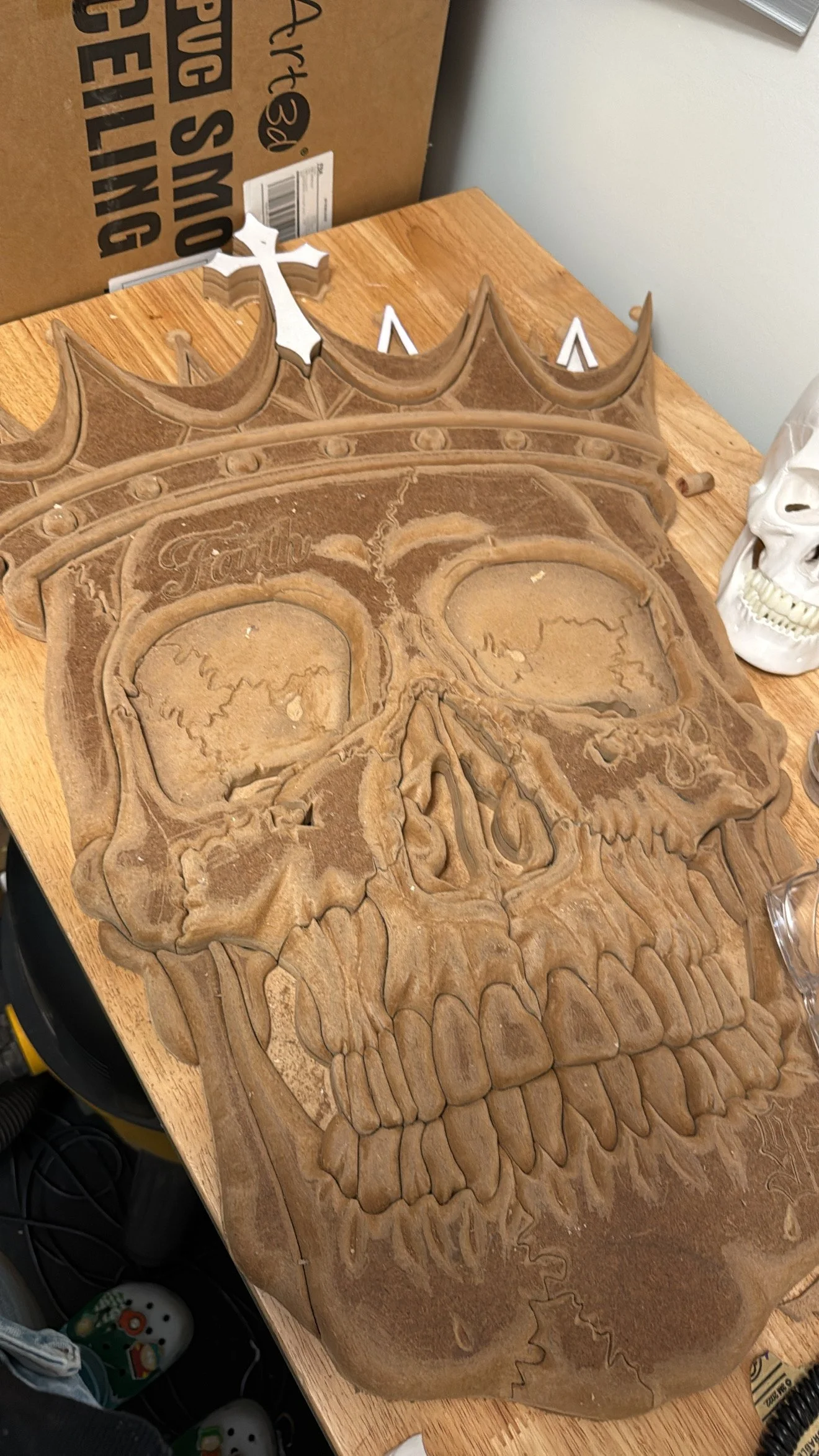 A 3D wooden carving of a human skull with hollow eye sockets, nasal cavity, and teeth, placed on a wooden surface.