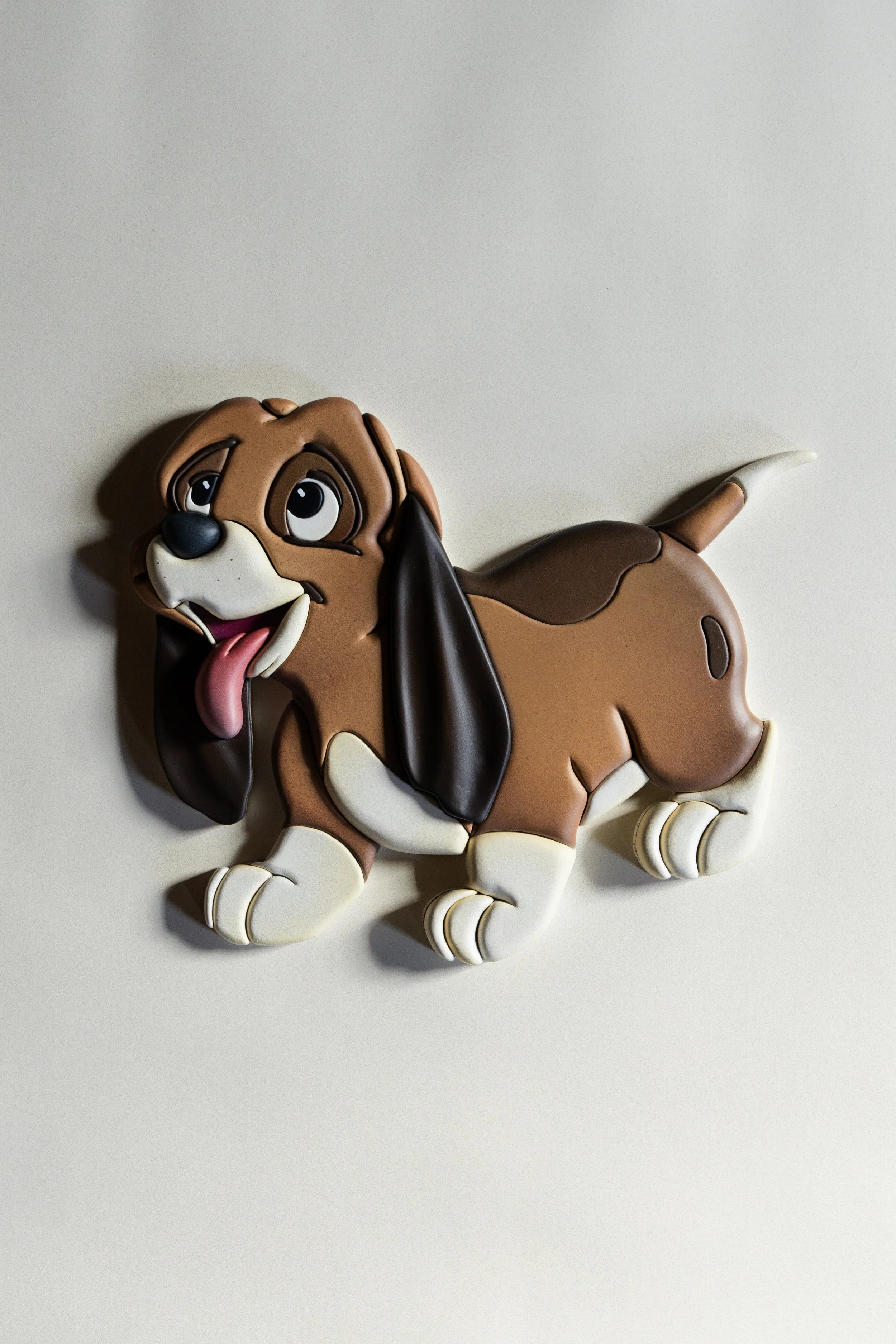 A large, colorful cookie shaped like a playful dog with a brown body, floppy ears, a pink tongue, and big expressive eyes, lying against a plain light background.