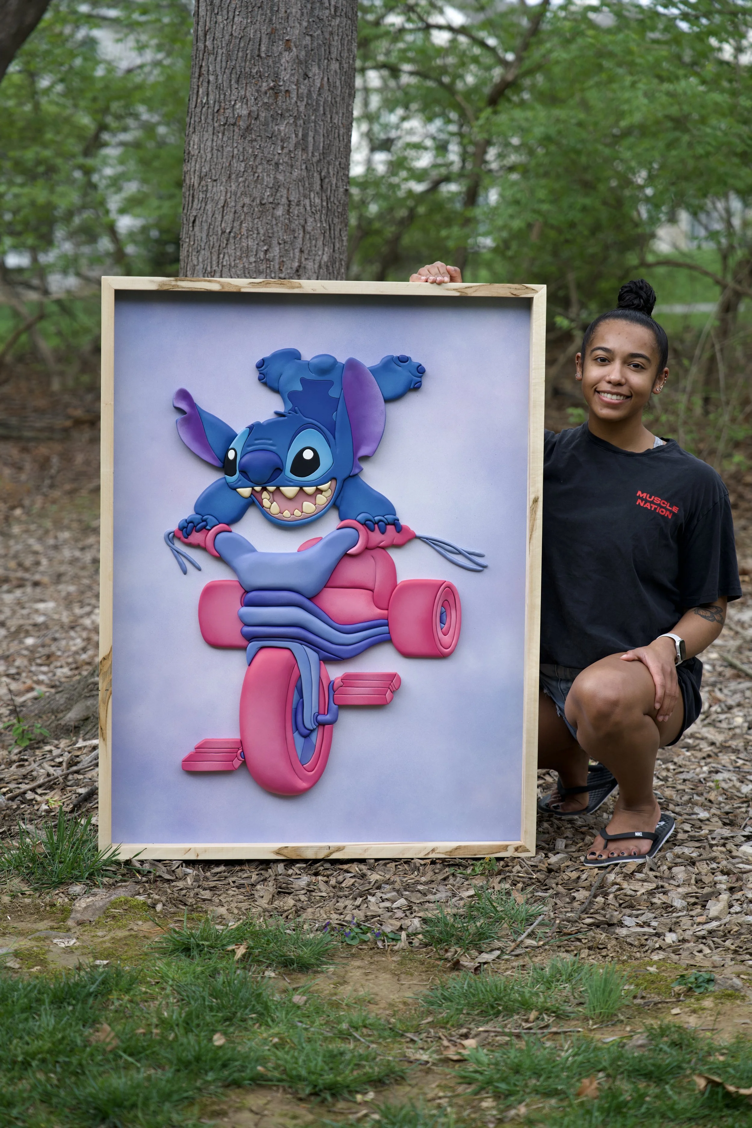 A smiling young woman crouching beside a large framed artwork of Lilo and Stitch character Stitch on a pink bicycle, outdoors in a wooded area with trees and green foliage in the background.
