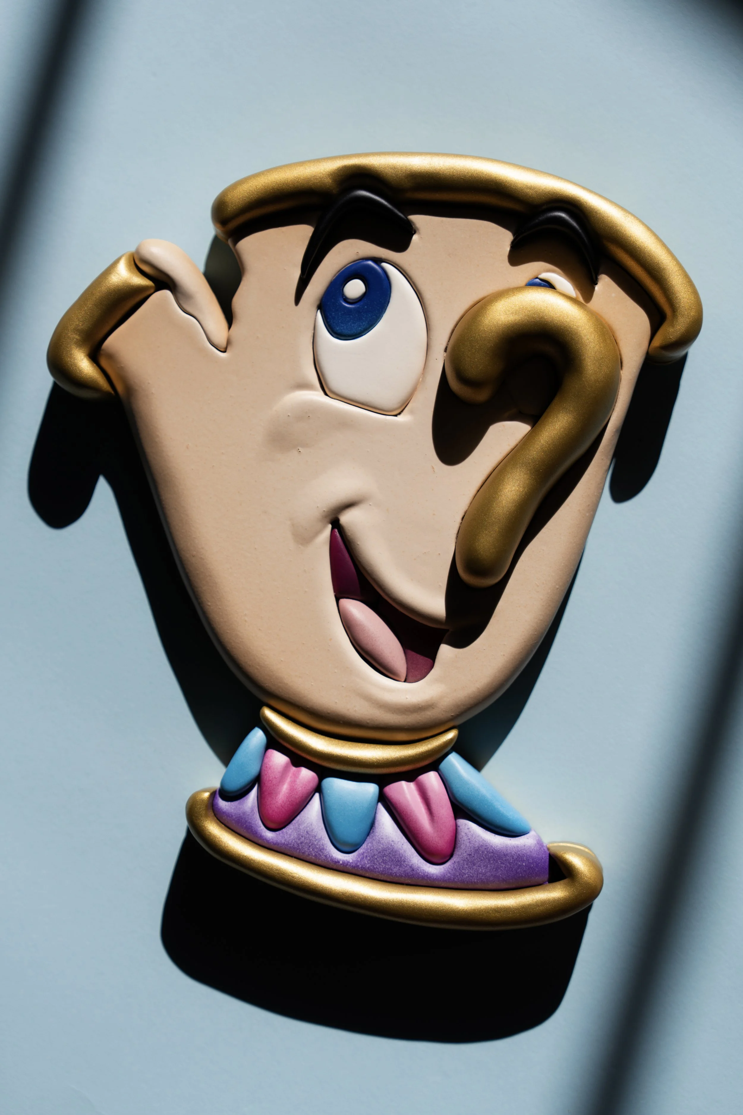 Close-up of a colorful cartoon character mask with exaggerated facial features, including large blue eyes, black eyebrows, and an open mouth, set against a light blue background. Chip wood art from Beauty and the beast