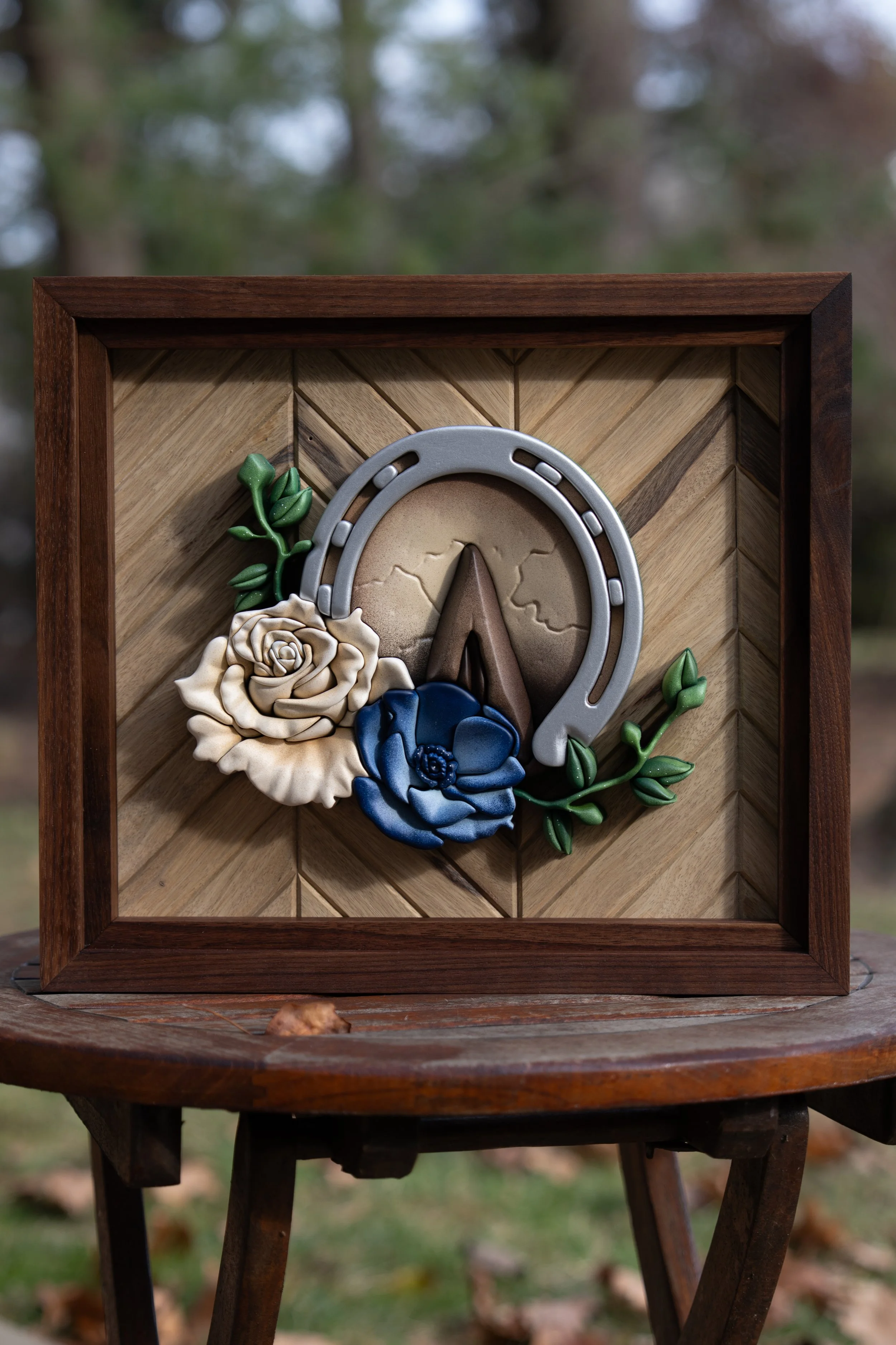 A decorative wall art piece featuring a horseshoe, roses, and greenery mounted on a wooden frame with a chevron pattern, placed on a wooden table outdoors.