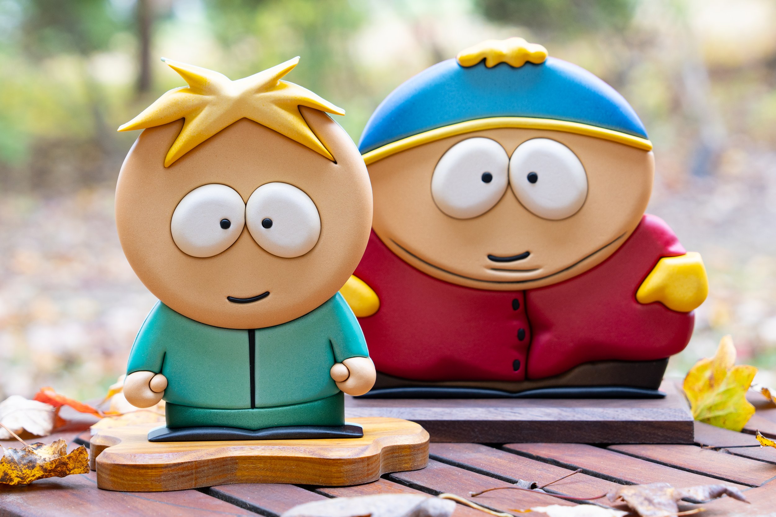 Colorful cartoon character figurines of Butters and Cartman on a wooden table outdoors, surrounded by autumn leaves, with a blurred natural background.