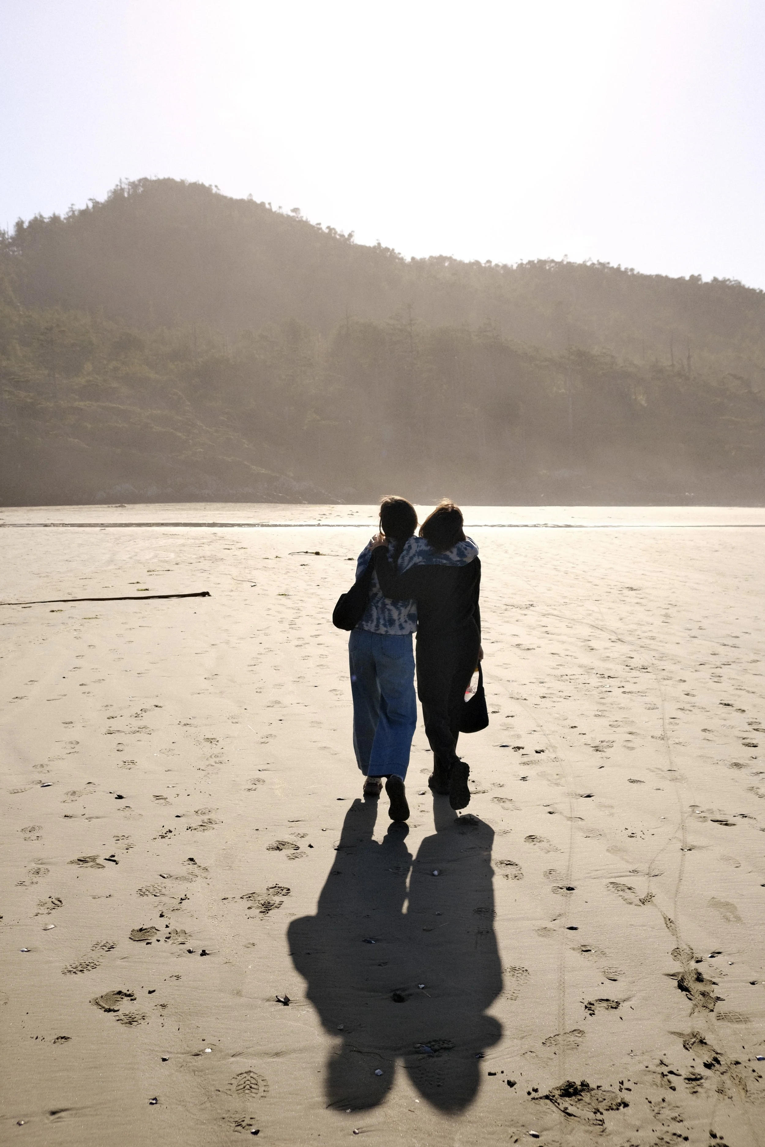 Two people walking on a sandy beach near water with hills and trees in the background on a sunny day.