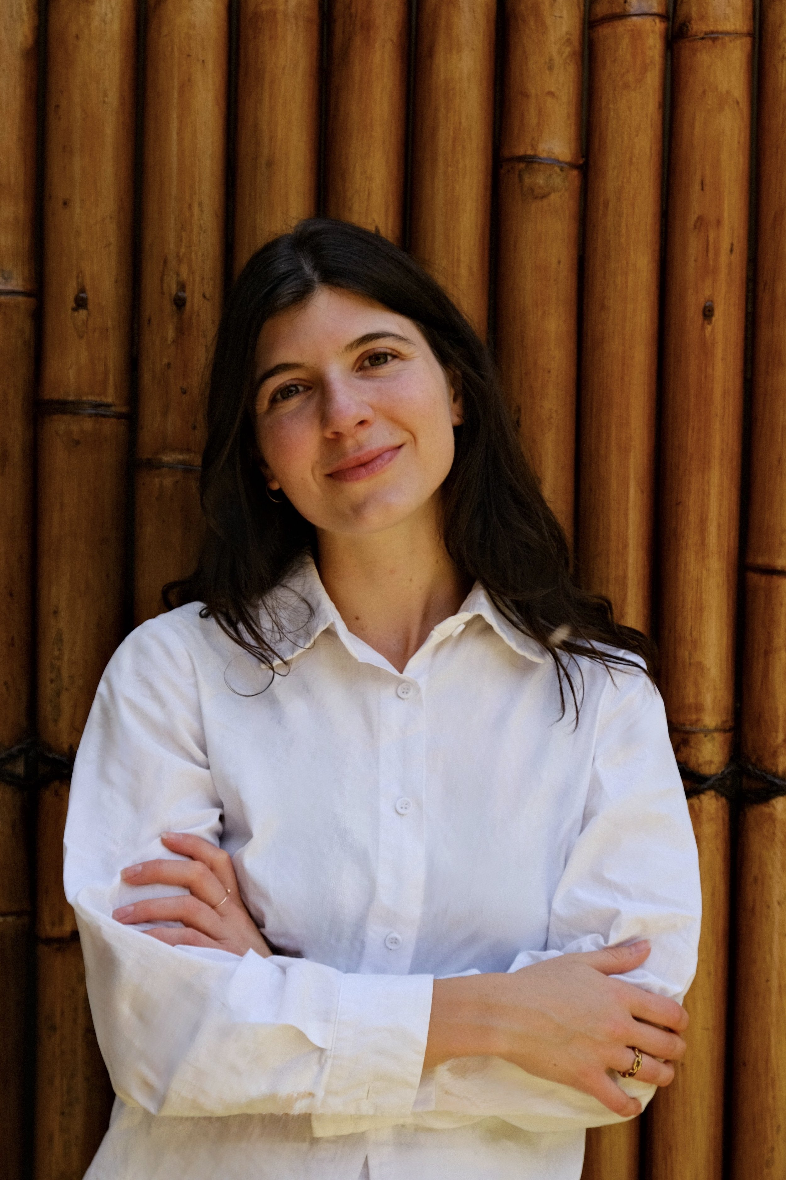 A woman with dark hair and fair skin, smiling slightly, wearing a white button-up shirt, stands with arms crossed in front of a bamboo wall.