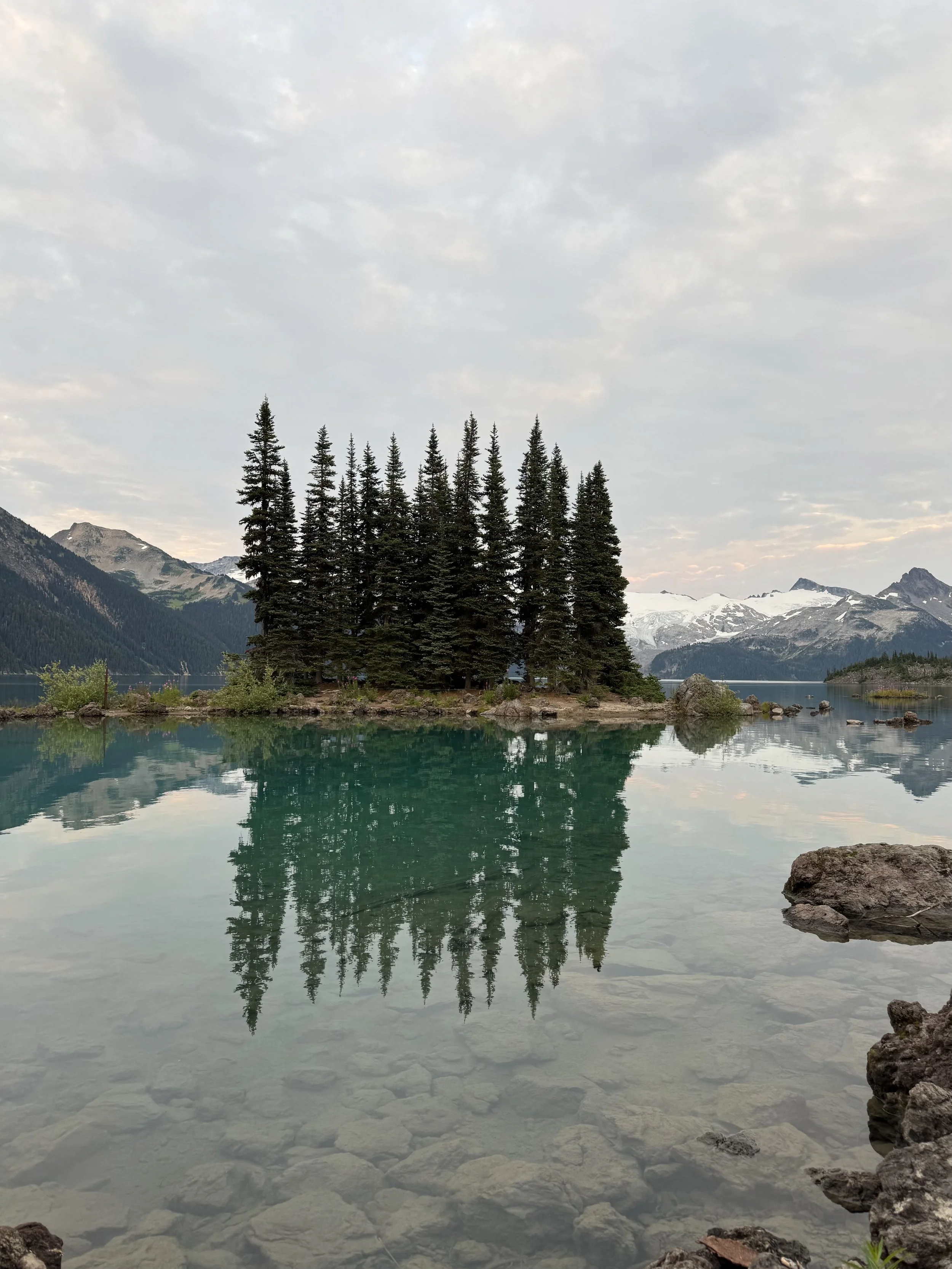 A tranquil lake with clear water reflecting horizon and trees, surrounded by mountains and overcast sky.