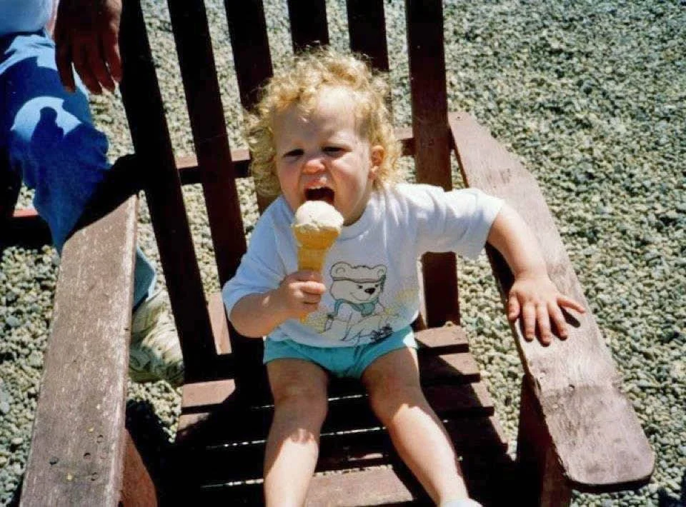 A young child with curly blonde hair sitting in a wooden chair outdoors, holding and about to eat an ice cream cone.