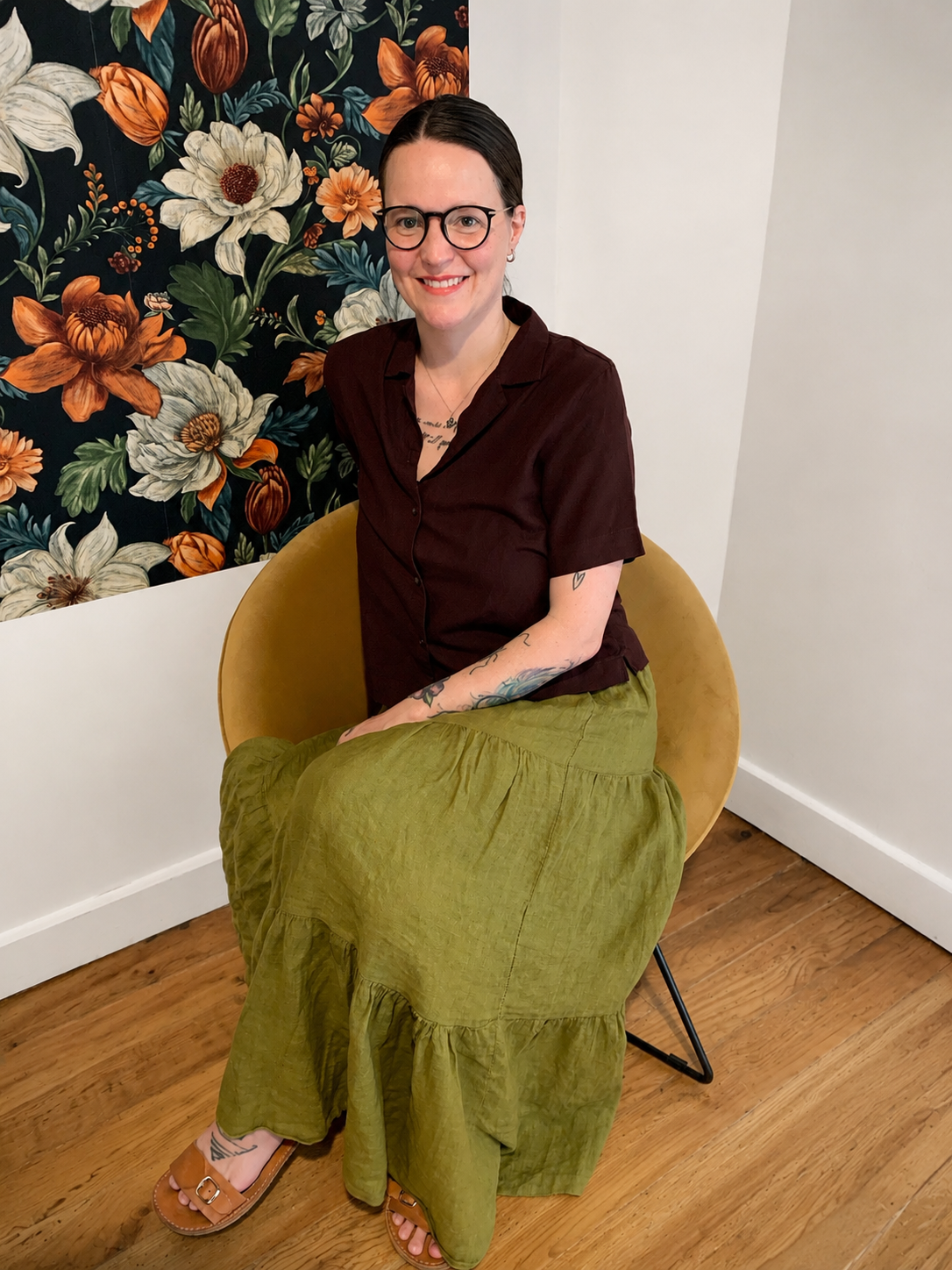 A woman with brown hair pulled back and glasses sitting on a yellow chair with a floral patterned wall behind her. She is smiling and wearing a maroon short-sleeved shirt, a green maxi skirt, and tan sandals, with tattoos on her arms and legs.