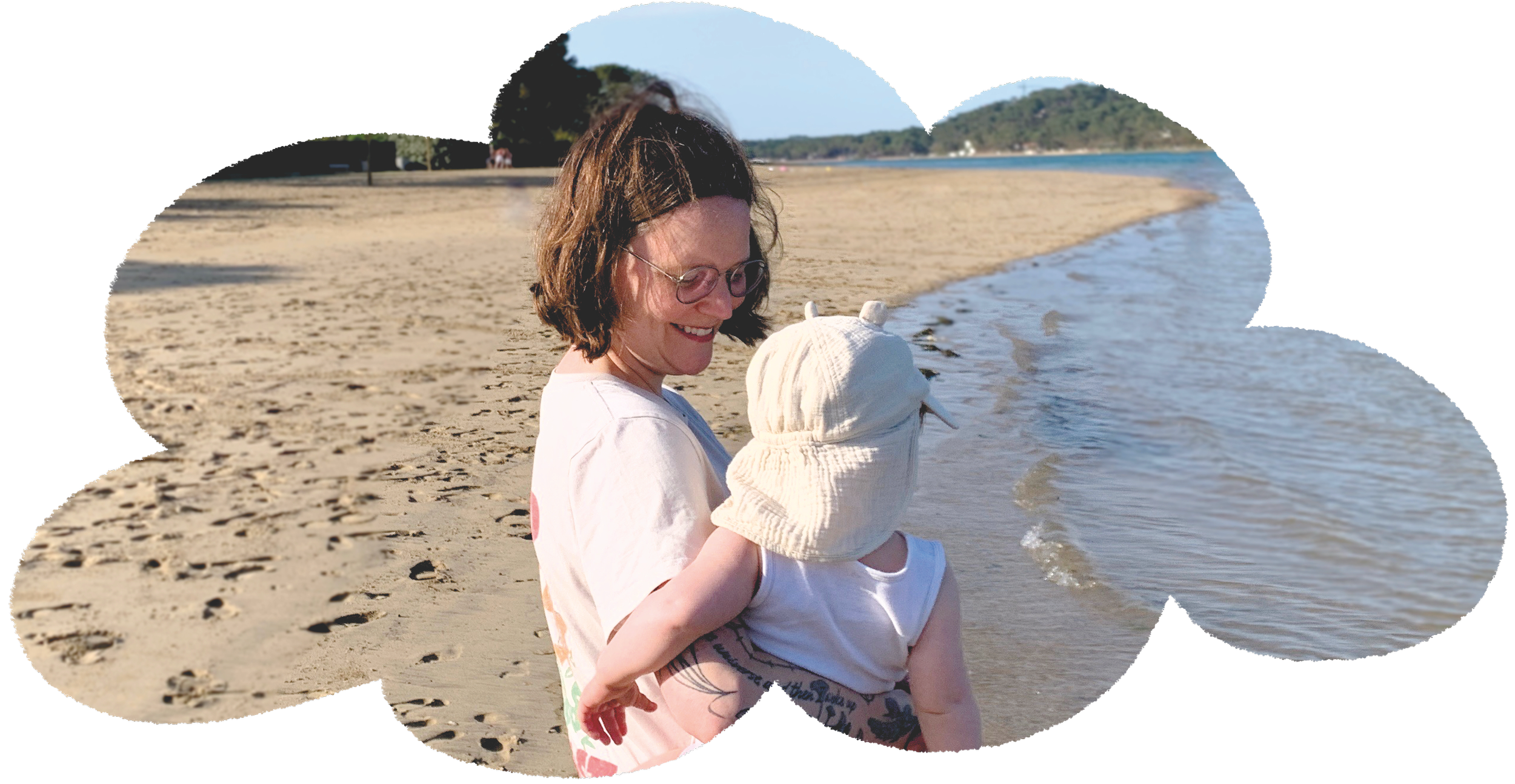 A woman holding a child at the beach with sand, water, and trees, in the background.