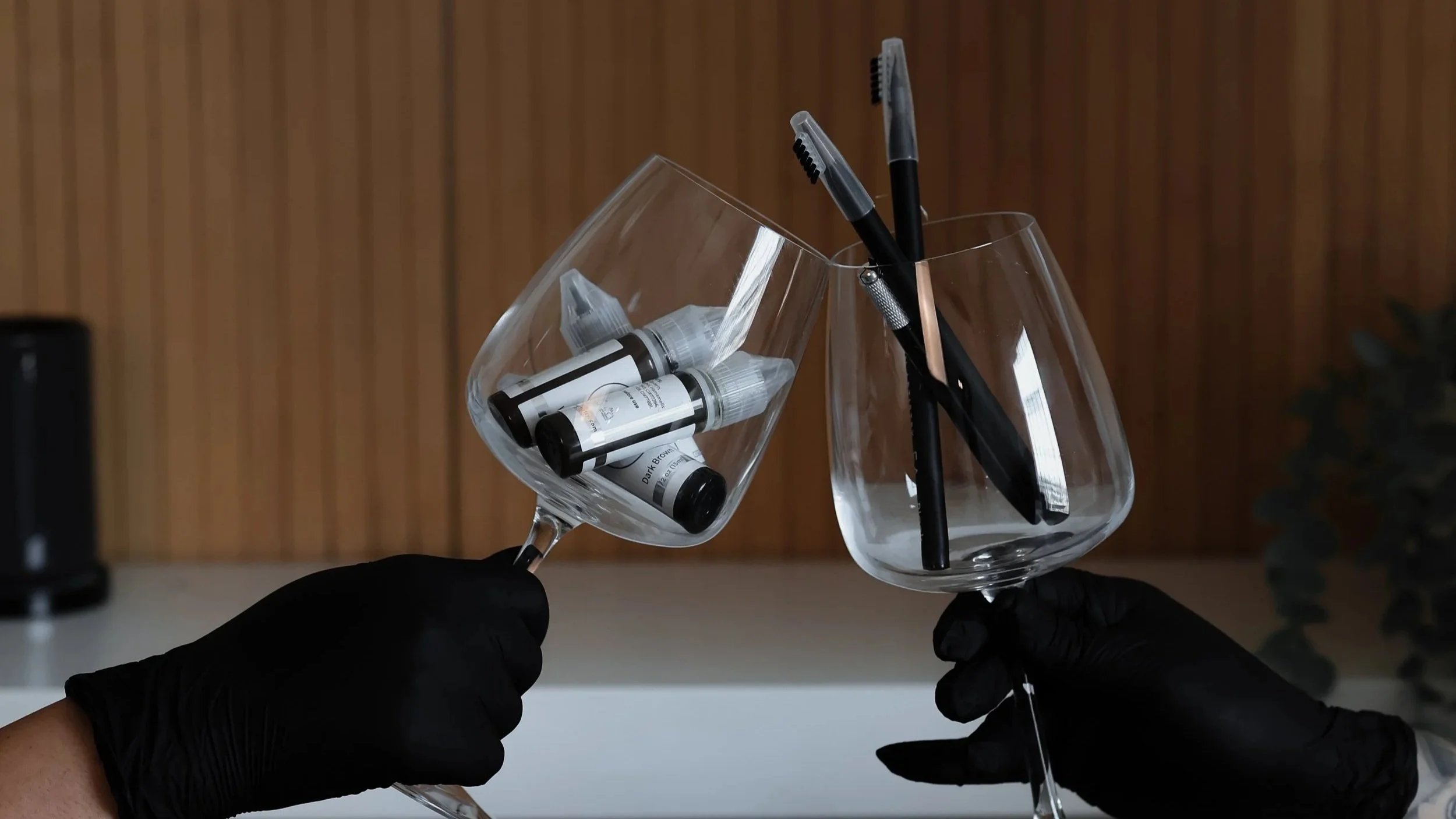 Moody editorial beauty product photo of black-gloved hands toasting wine glasses filled with serum vials and brow pencils