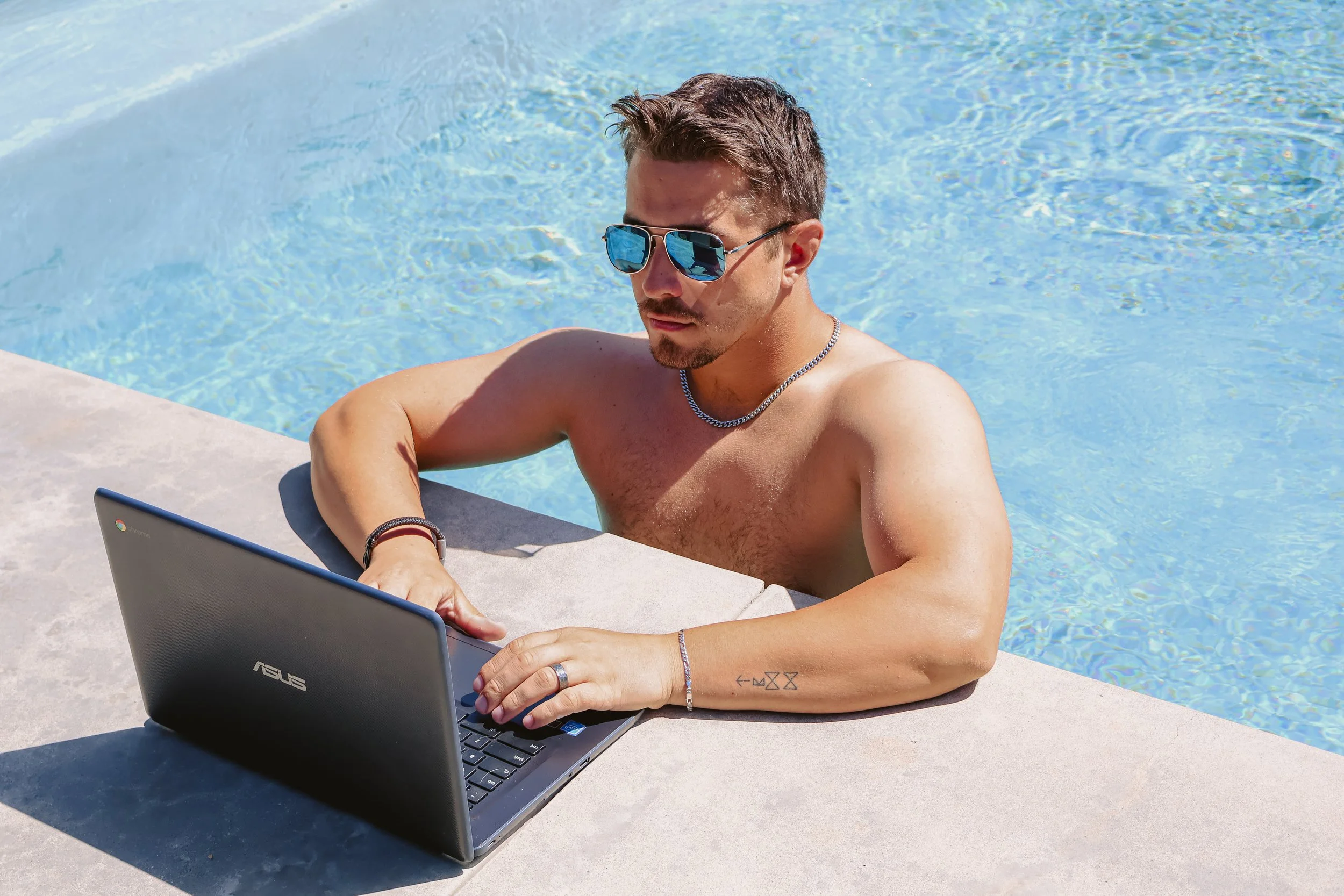 Man in sunglasses working on a laptop at the edge of a pool, lifestyle brand photography by Poppy House Media