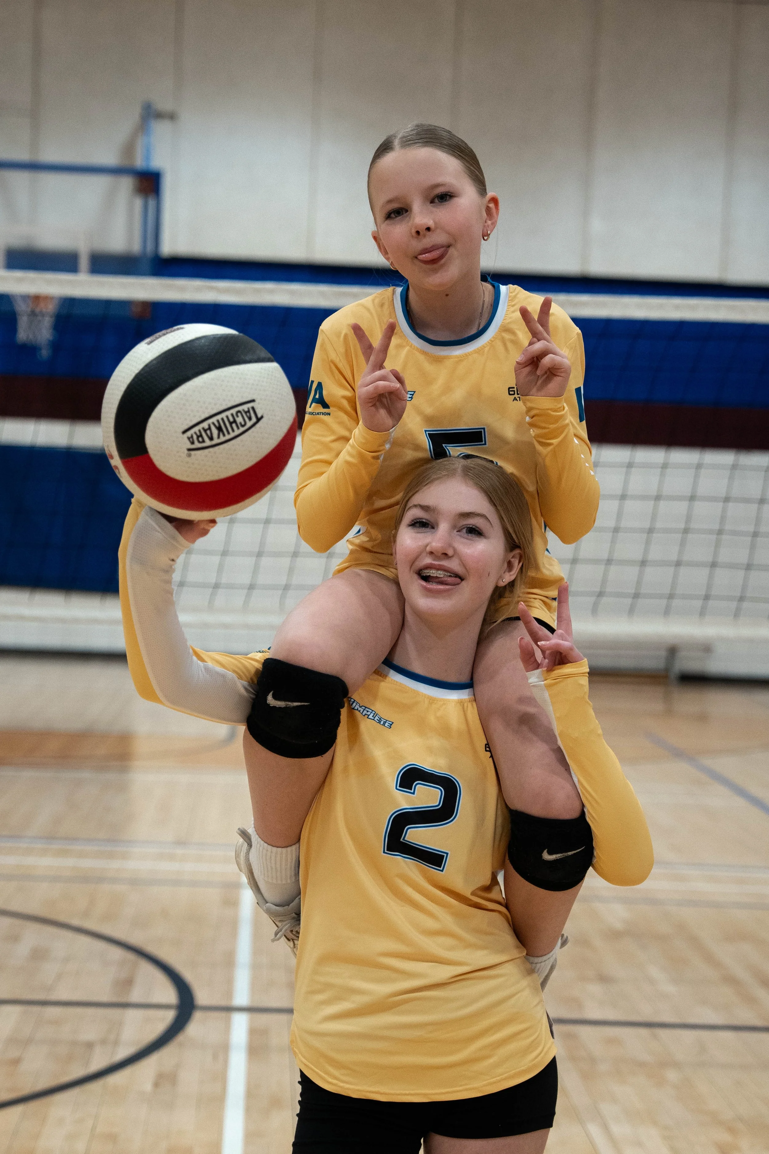 Two young women in yellow volleyball uniforms inside a gymnasium, making playful gestures with their tongues out. One is sitting on the other's shoulders, holding an AiChihara volleyball. They are in front of a volleyball net and court.