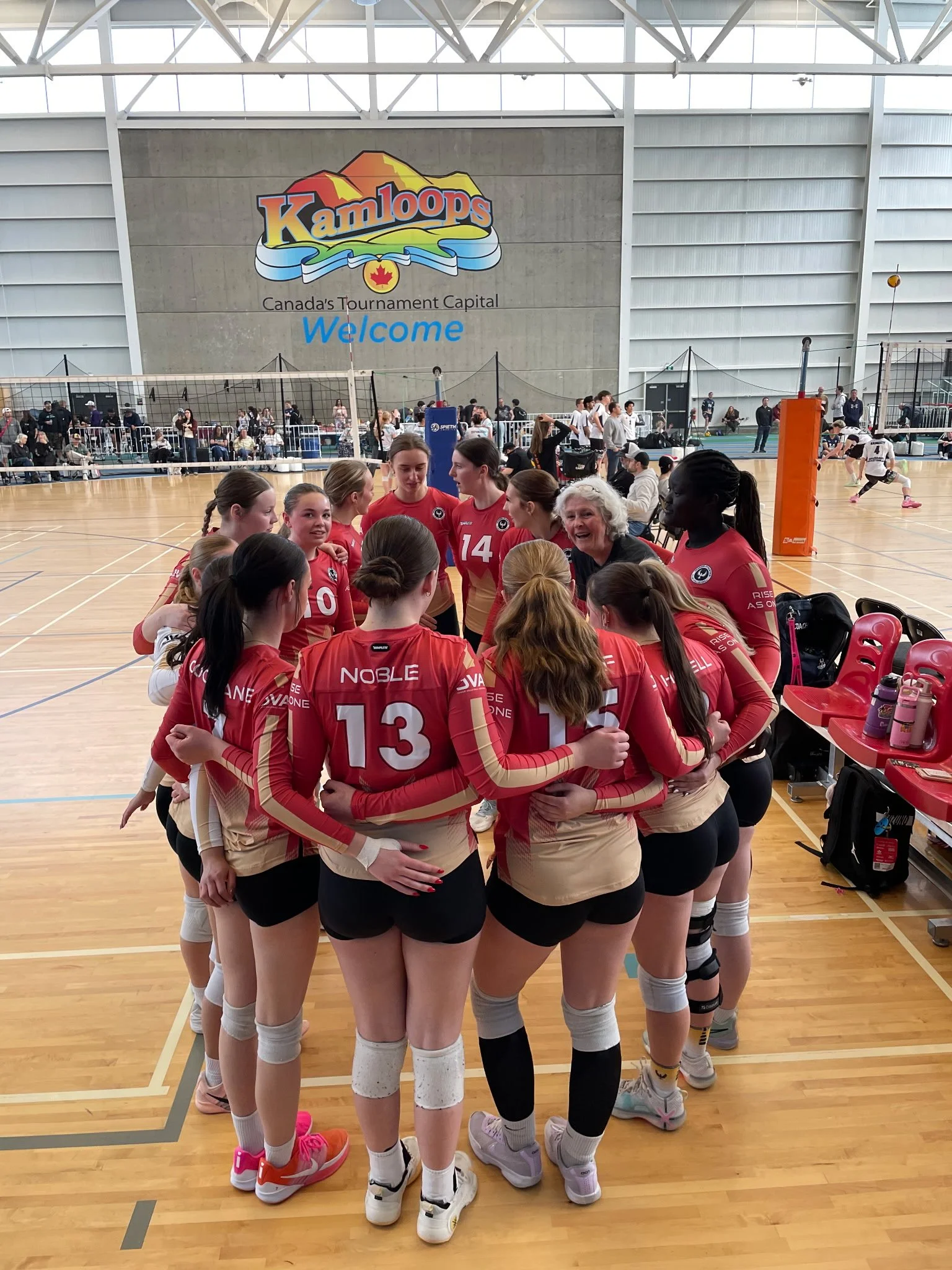 A volleyball team huddle with players wearing red and beige uniforms inside a gymnasium with a wooden floor, with a large Kamloops sign and volleyball courts in the background.