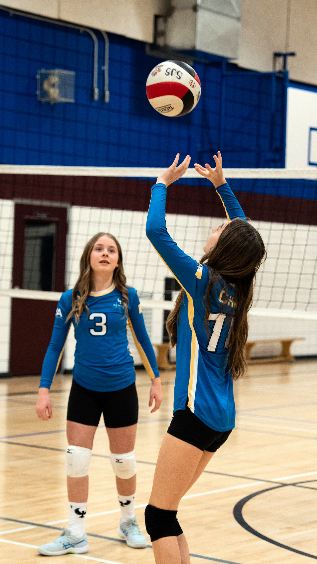 Two girls playing volleyball indoors, one is setting the ball while the other watches, both wearing blue jerseys with yellow accents and black shorts.