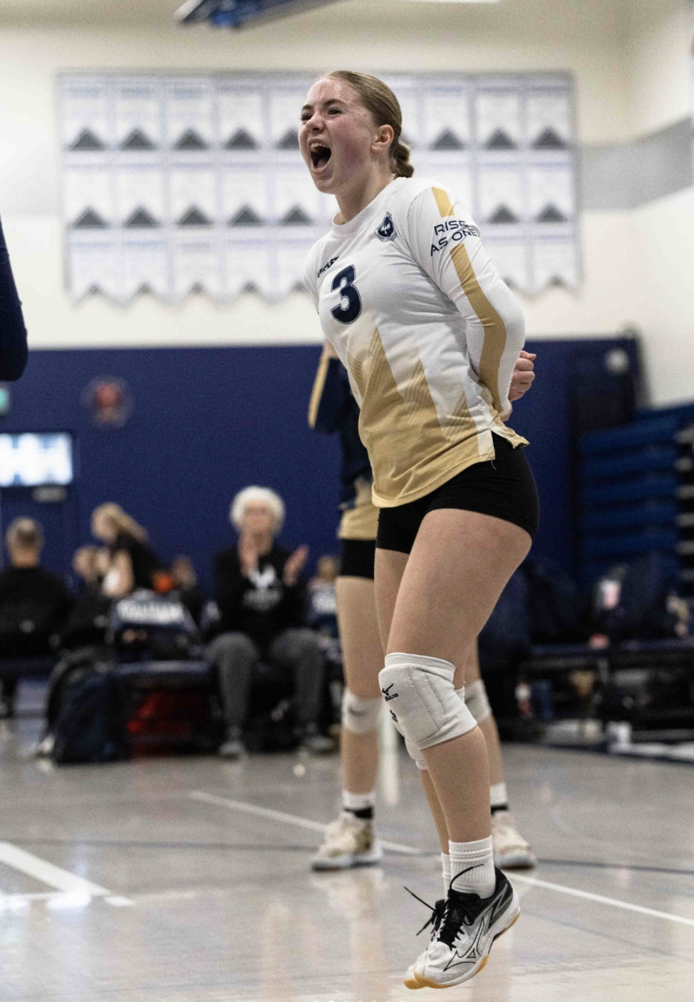 A female volleyball player wearing a white jersey with the number 3, black shorts, and knee pads, jumping with her mouth open in celebration during a match.