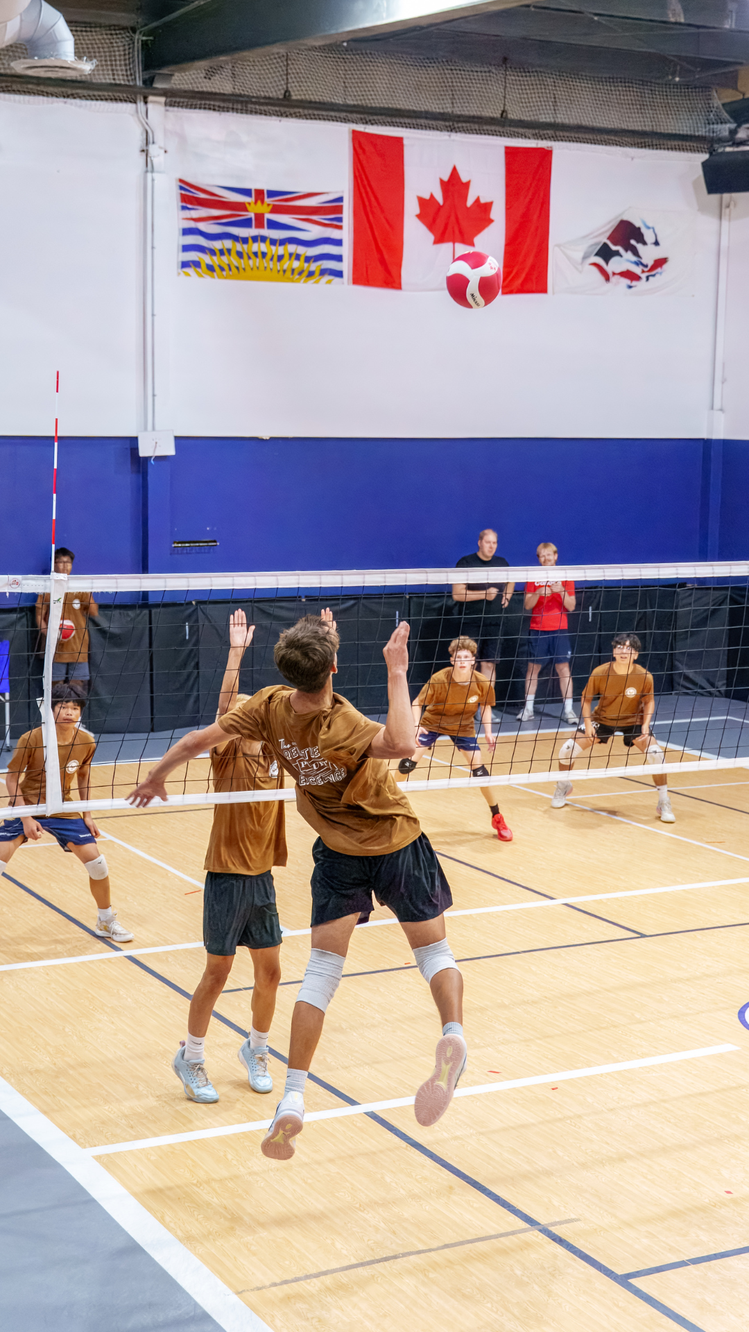 Young boys playing indoor volleyball, with a player jumping to hit the ball over the net, and a volleyball in midair. Background features flags, including Canada, a British Columbia flag, and a stylized mountain and whale flag.