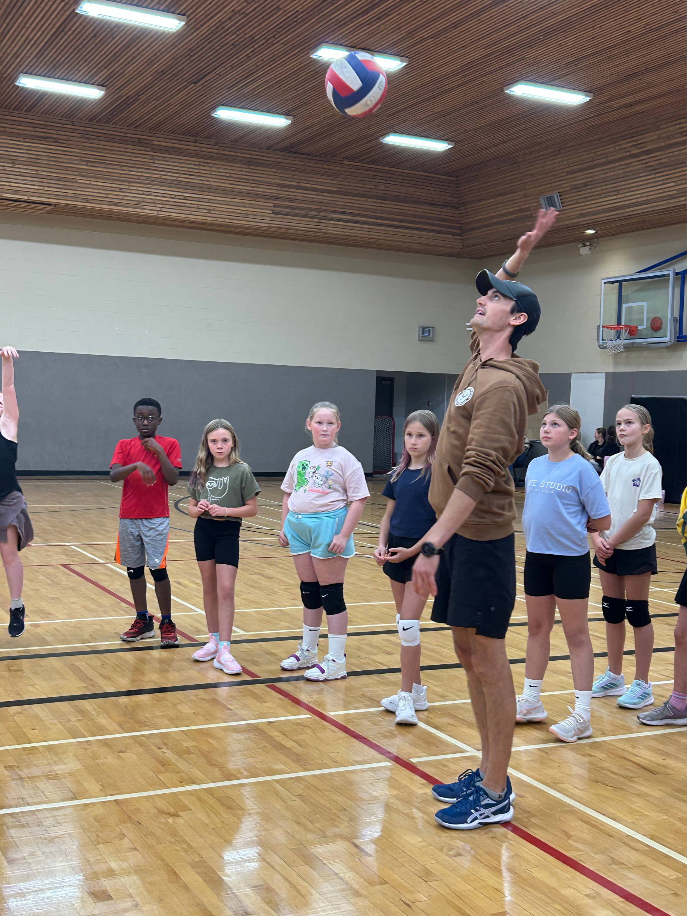 A group of children and a coach are in a gymnasium practicing volleyball. The coach is hitting a volleyball in the air. Volleyball Kelowna Genesis Athletics Club