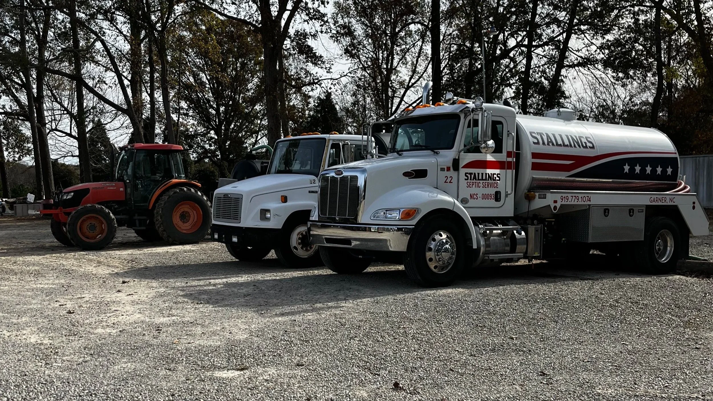 A septic service truck parked next to a red tractor, with trees in the background.