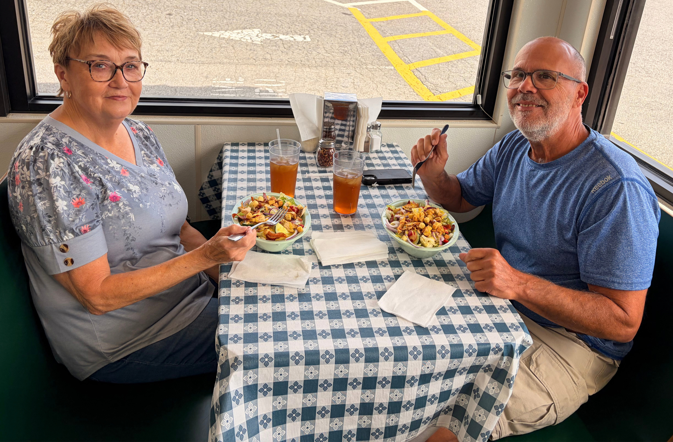 A smiling woman with short blonde hair and glasses wearing a gray and pink floral shirt, sitting at a table with a man who has a gray beard and glasses, wearing a blue t-shirt. They are eating salads with drinks inside of the restaurant.