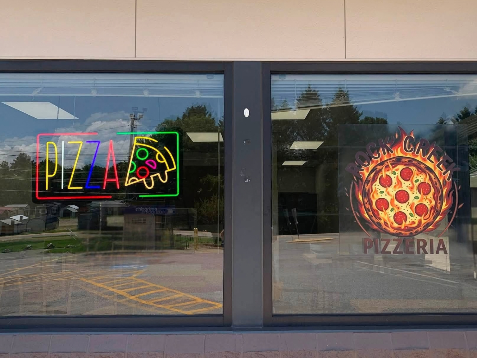 Neon signs on a restaurant window, one with the word 'PIZZA' and a pizza slice, and the other with a pizza surrounded by flames and the words 'ROCK CREEK PIZZERIA'.
