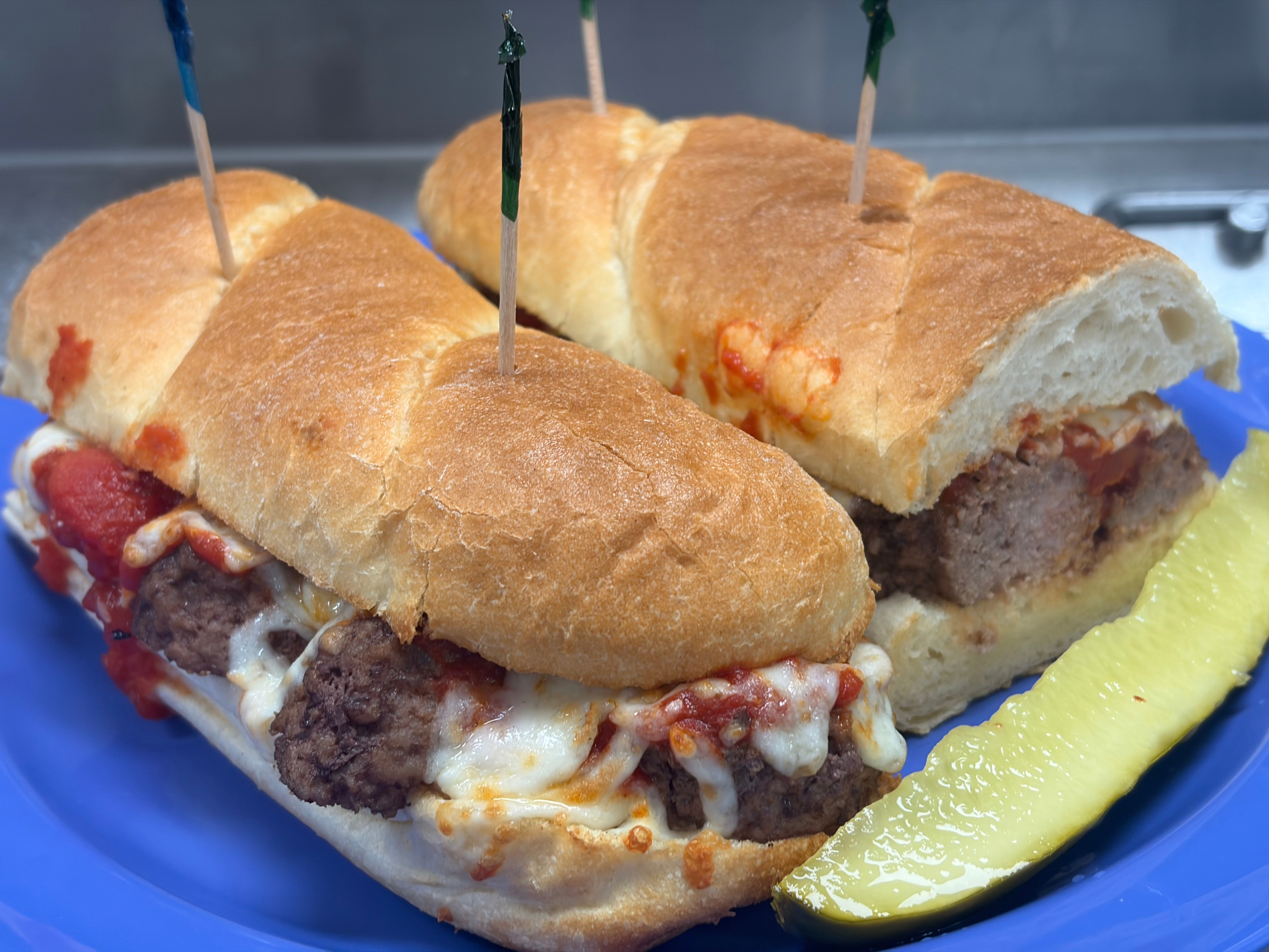 A plate with a meatball sub sandwich topped with melted cheese and tomato sauce, served with a pickle on the side.