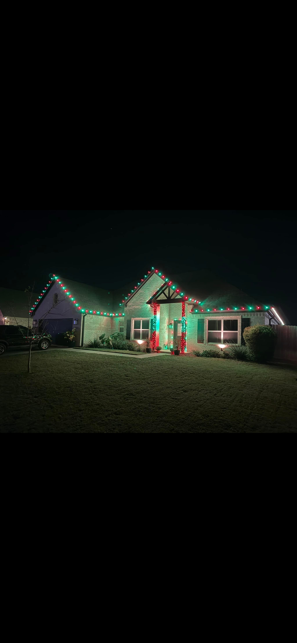 A single-story house decorated with red and green Christmas lights at night. The house has a front porch with two columns wrapped in red lights, two windows with lighting at the base, and a small tree in the yard.