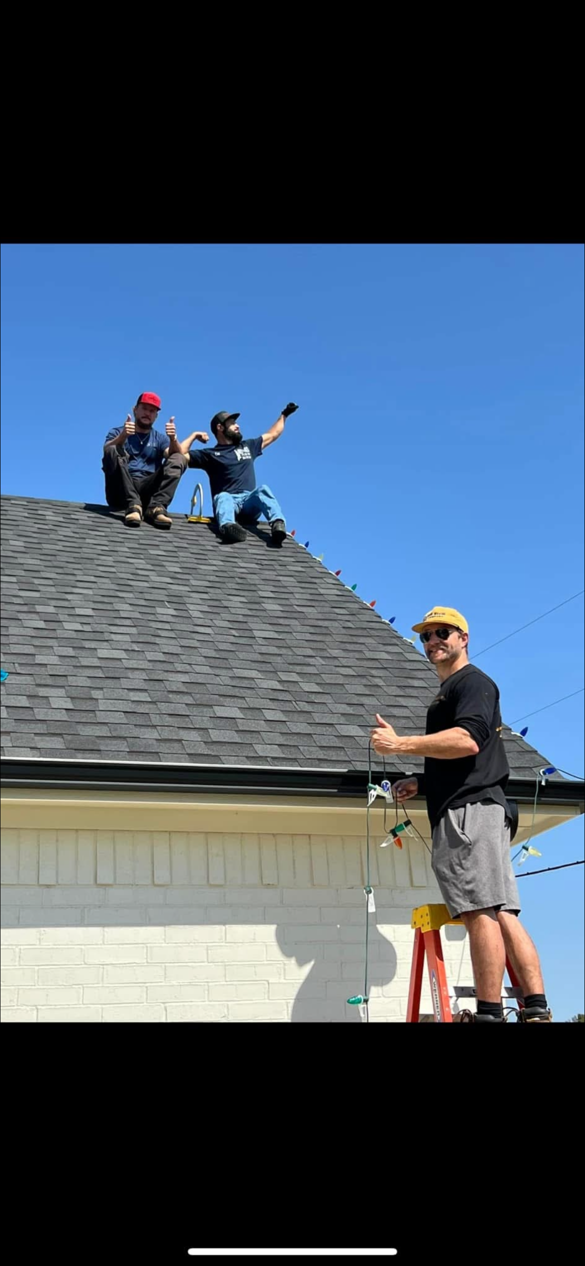 Three men on a house roof and one man on a ladder with Christmas lights, with a clear blue sky background.