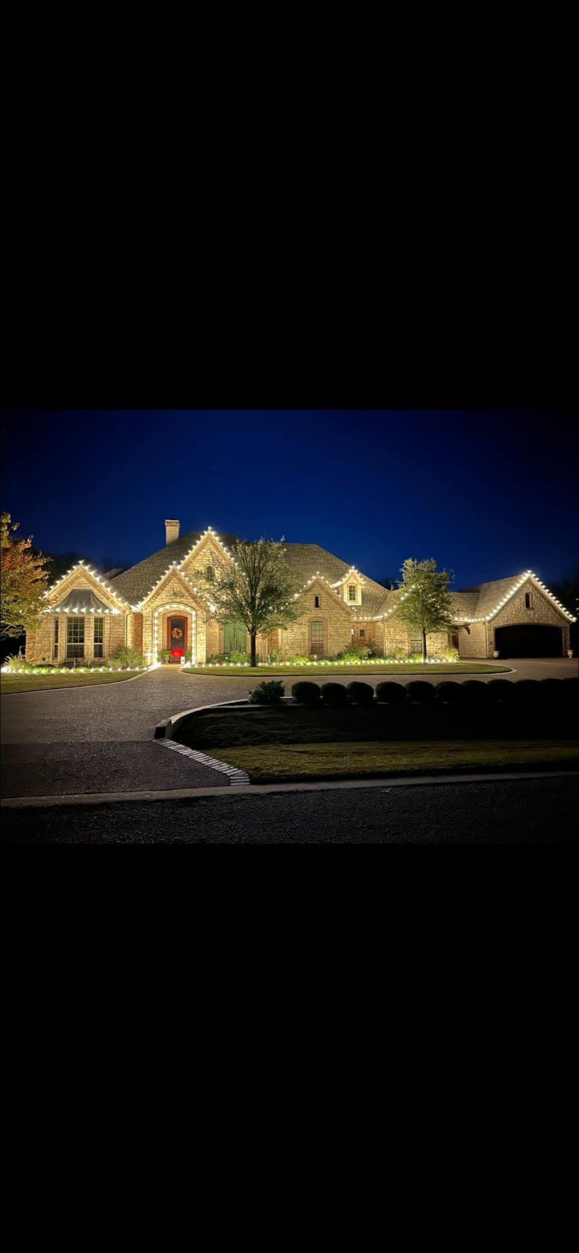Brightly lit brick house at night with festive holiday lights outlining roof and windows, surrounded by a driveway, trees, and landscaped yard.