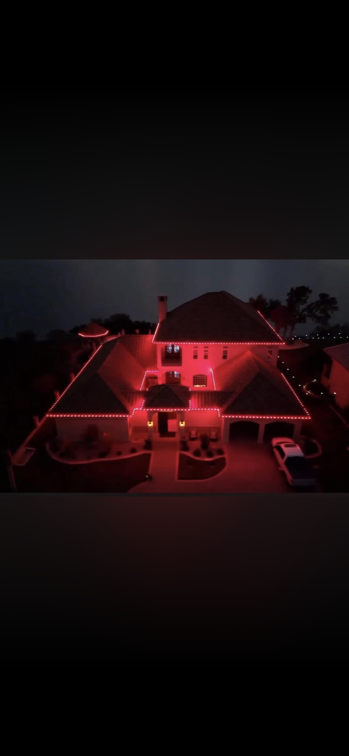 A large house decorated with red string lights outlining the roof and architectural features, with a driveway, landscaped yard, and parked white car visible in the front.