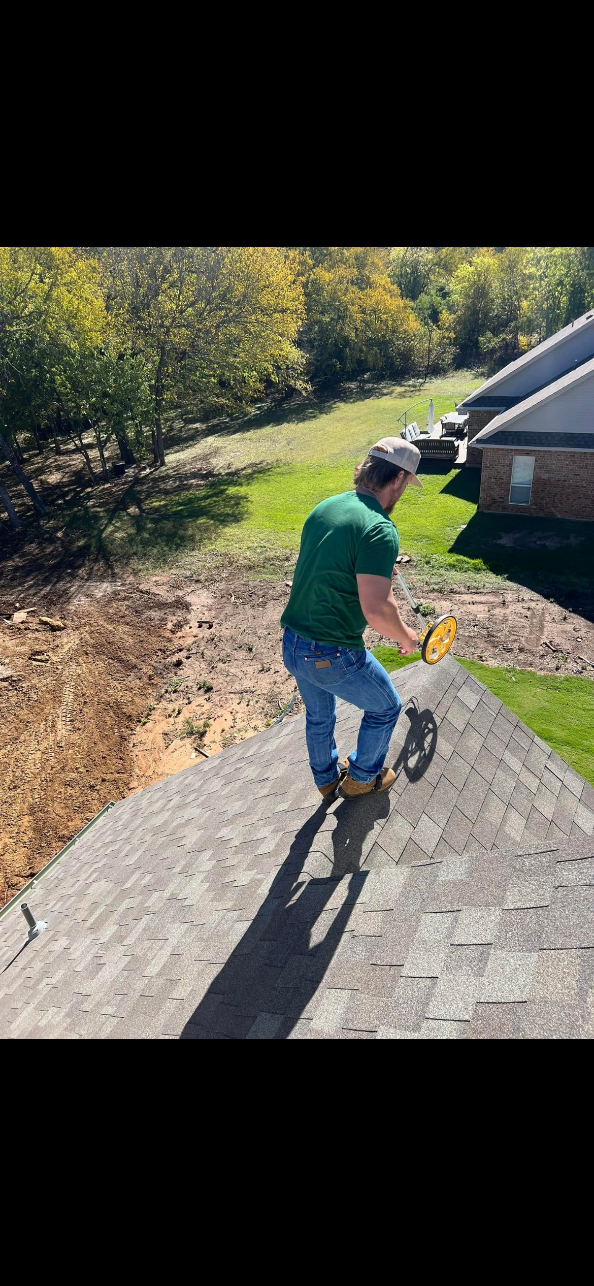 A man on a roof using a tool, with a backyard and house visible in the background, surrounded by trees on a sunny day.