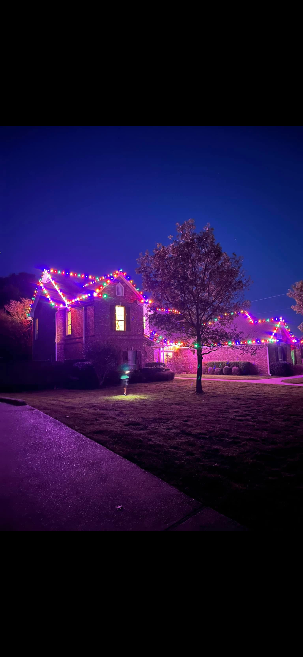 A two-story house decorated with colorful Christmas lights at night, with a tree in the front yard and a person holding a lantern walking on the sidewalk.