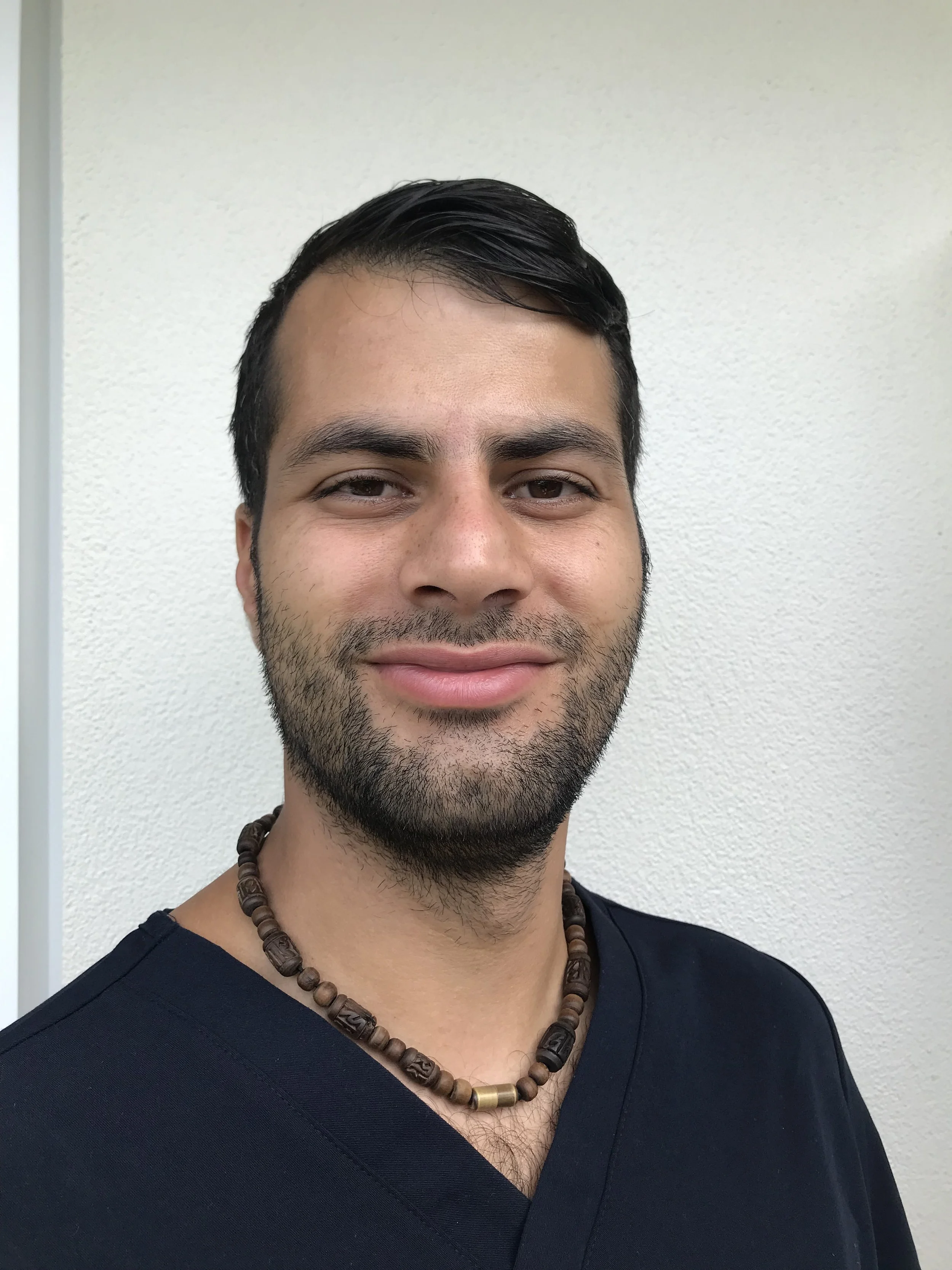 Close-up of a smiling man with short black hair, light brown skin, wearing a black shirt and a beaded necklace, standing against a plain off-white wall.