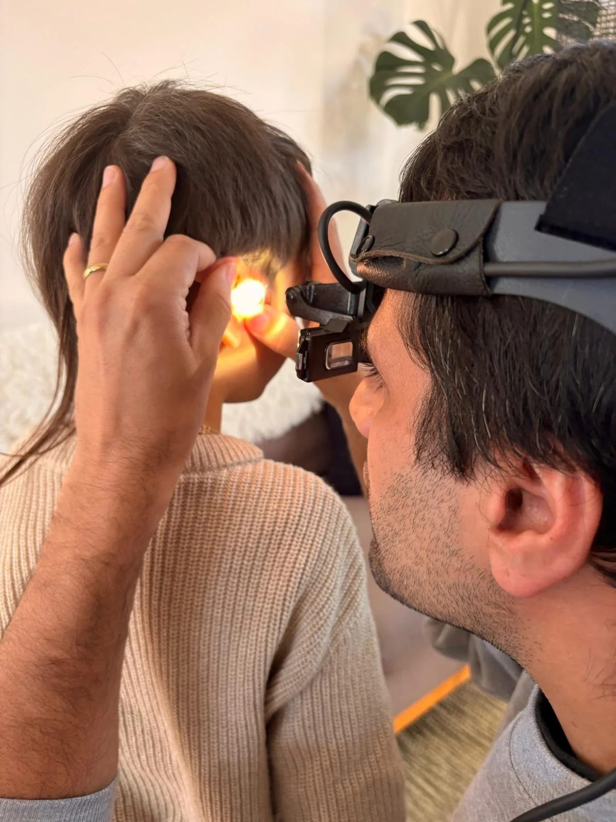 A medical professional using an otoscope to examine a child's ear. The child has dark hair and is wearing a beige sweater. The professional is wearing glasses and a headlamp for better visibility.