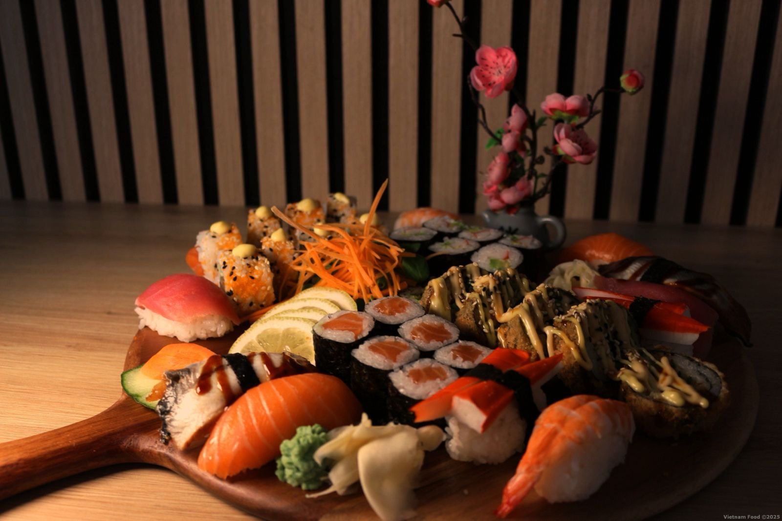 Assorted sushi platter with various types of sushi, sashimi, lemon slices, pickled ginger, wasabi, and a flower arrangement on a wooden serving board.