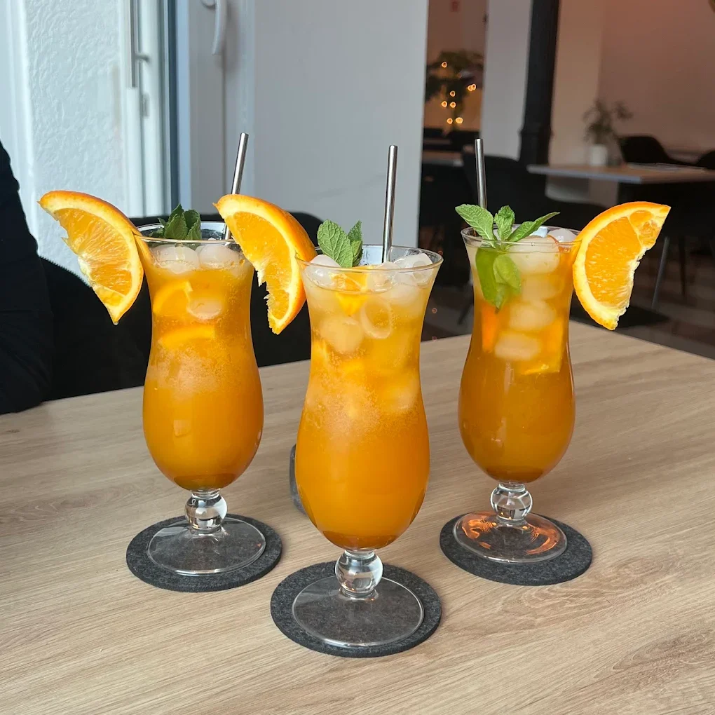 Three tall glasses of orange-colored cocktail with ice, orange slices, and mint leaves on a wooden table.
