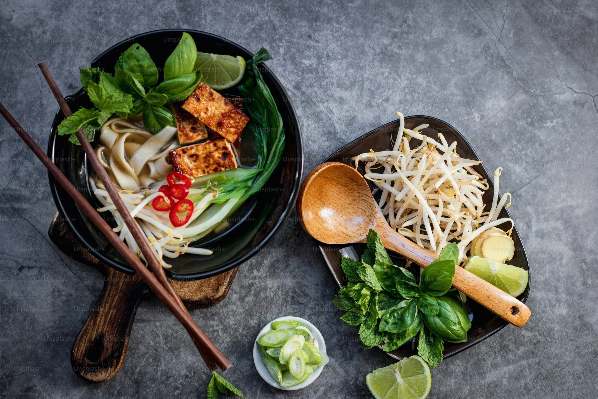 A bowl of Vietnamese noodle soup with fresh herbs, lime, sliced chili peppers, and grilled tofu, accompanied by a side of bean sprouts, lime wedges, and herbs on a dark surface.