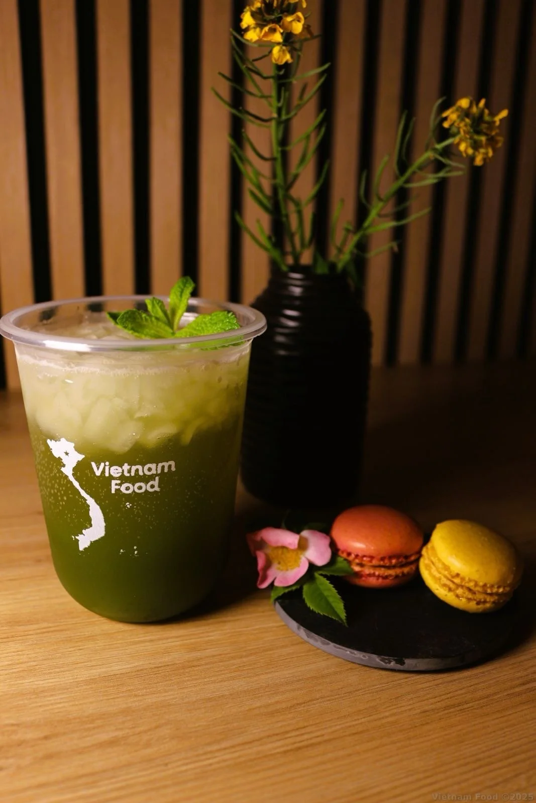 A Vietnamese food drink in a clear cup with mint garnish, with a black vase of yellow flowers in the background. A small plate with pink and yellow macarons and a pink flower is also on the table.