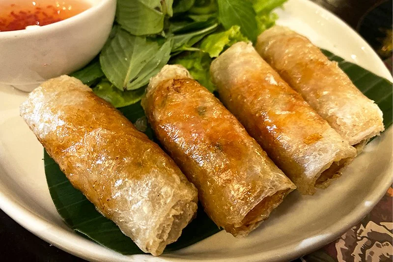 Four fried spring rolls on a white plate with lettuce and a small bowl of dipping sauce.