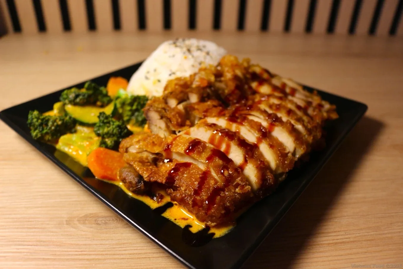 Plate of food with grilled chicken topped with sauce, steamed mixed vegetables including broccoli and carrots, a scoop of rice, all served on a black square plate.