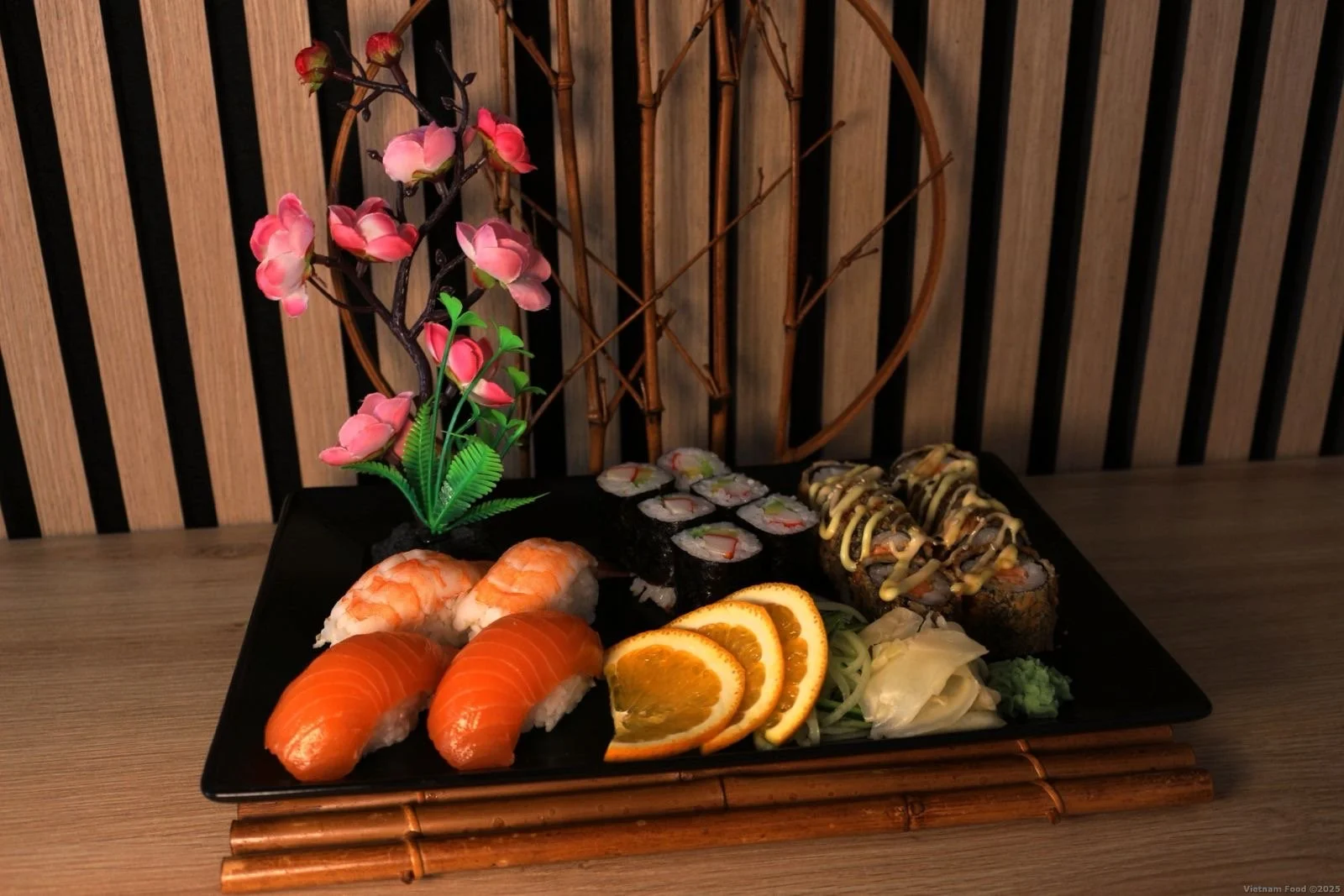 A black rectangular plate of assorted sushi with salmon, shrimp, sushi rolls, orange slices, ginger, wasabi, and garnishments, with decorative pink cherry blossom and bamboo accents in the background.