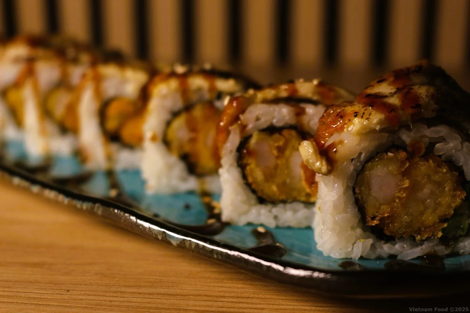 Close-up of a plate of sushi rolls with rice, tempura and eel, topped with eel sauce, placed on a wooden table.