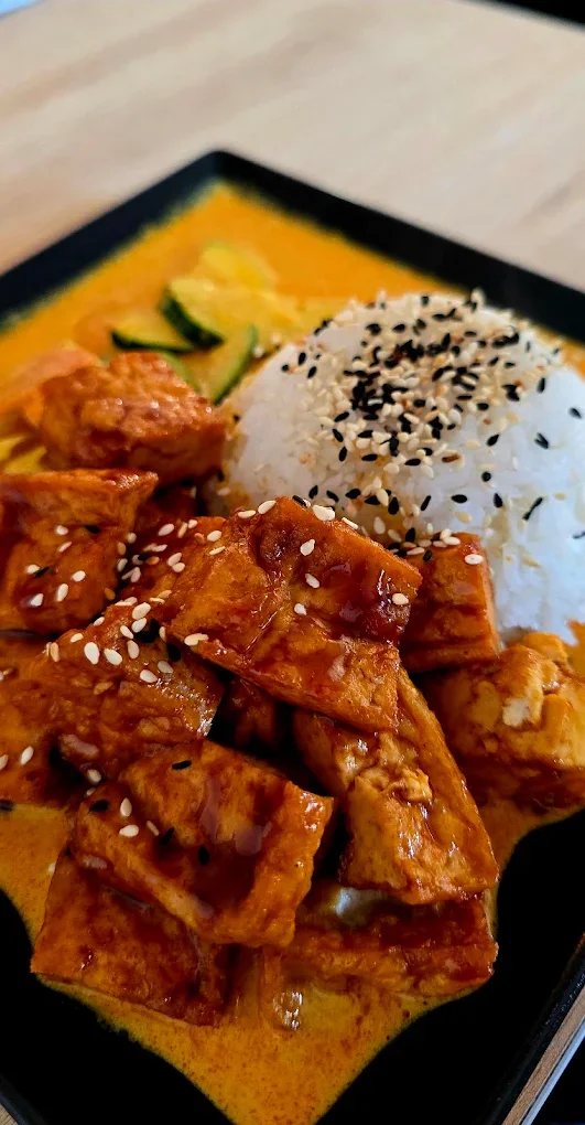 Asian-inspired dish on a black tray with rice topped with black and white sesame seeds, grilled chicken pieces glazed with sauce and sprinkled with sesame seeds, and slices of cucumber, served on a wooden table.