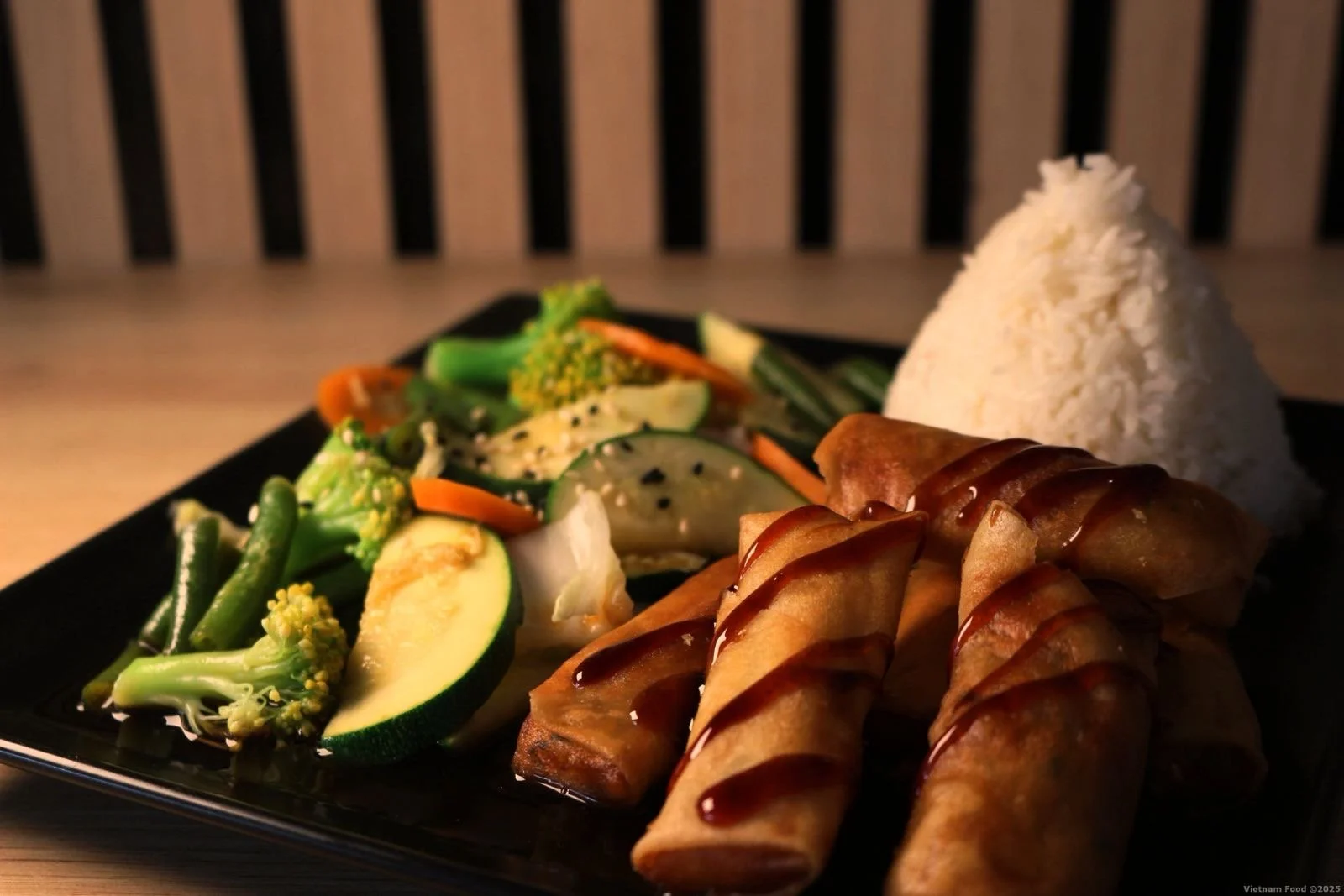 A black square plate with mixed vegetables, a serving of white rice, and four spring rolls drizzled with sauce.
