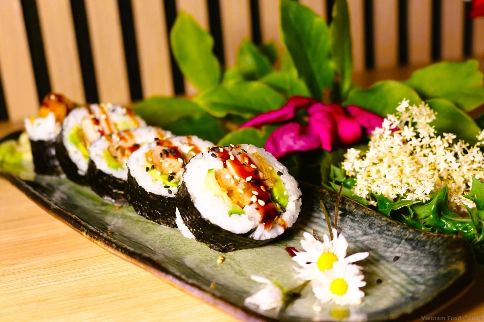 A plate of sushi rolls garnished with vegetables and sauces, with pink and white flowers and green leaves in the background.