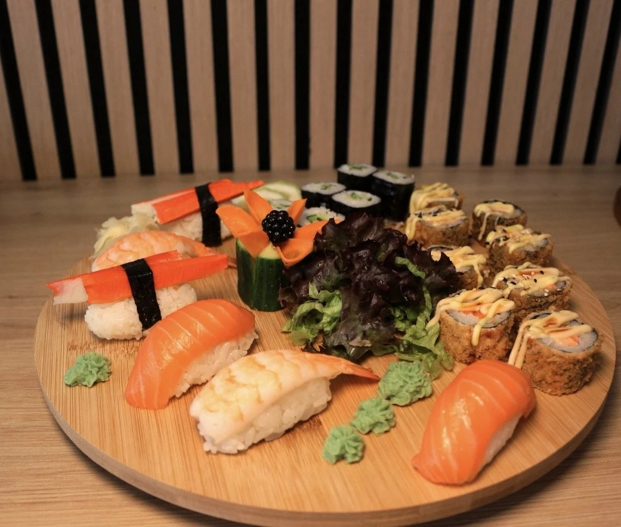 A wooden platter with assorted sushi pieces, including nigiri with salmon, shrimp, and crab, plus various sushi rolls garnished with sauce, on a wooden table with a striped background.