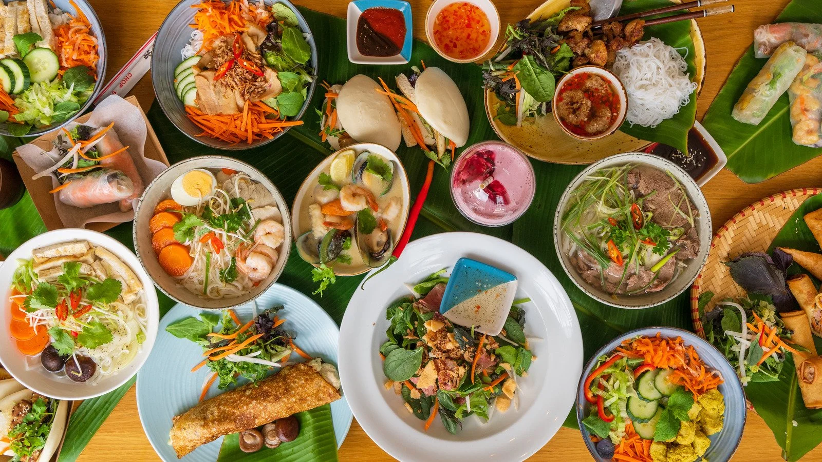 A variety of Vietnamese dishes on a wooden table. Includes salads, spring rolls, noodle bowls, and grilled meats served on banana leaves, along with dipping sauces and a colorful cocktail.