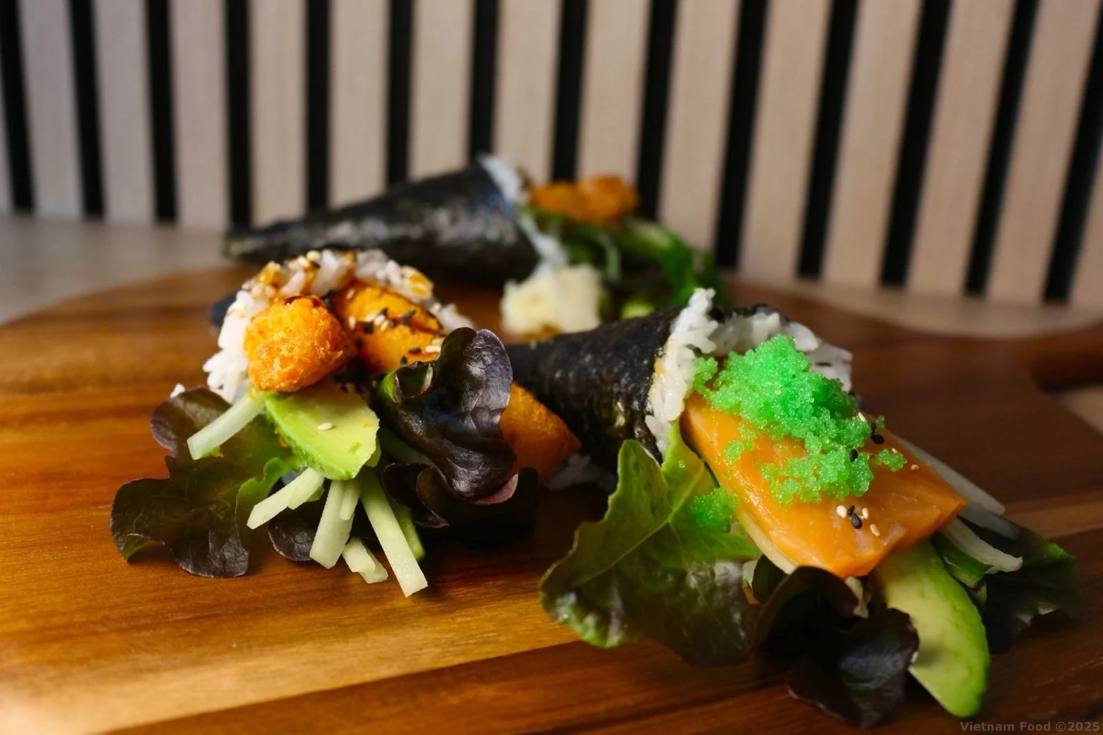 Sushi rolls with avocado, salmon, green roe, and mixed greens on a wooden serving board.