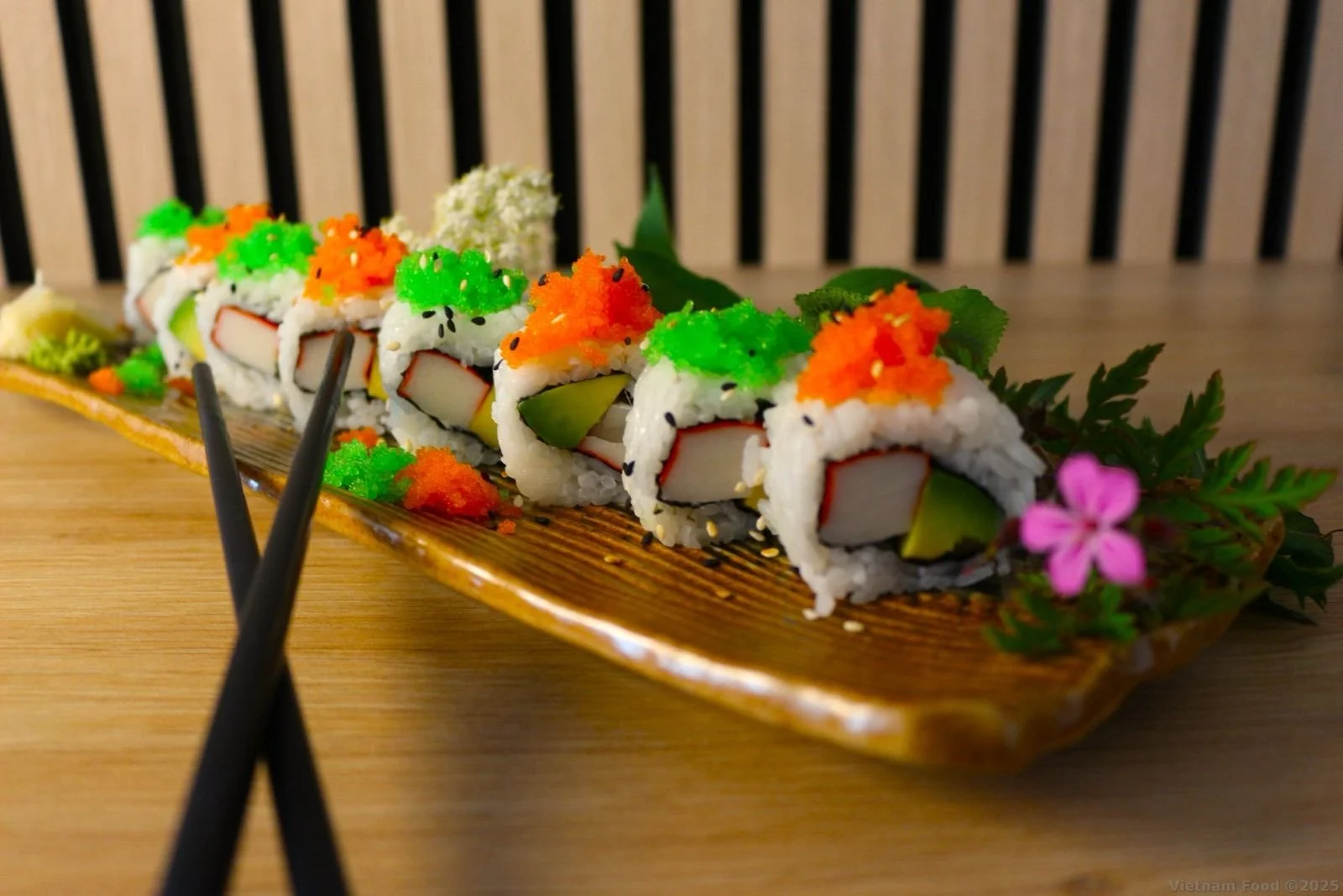 A sushi platter with colorful sushi rolls topped with green and orange fish roe, garnished with leaves and a pink flower on a wooden plate.