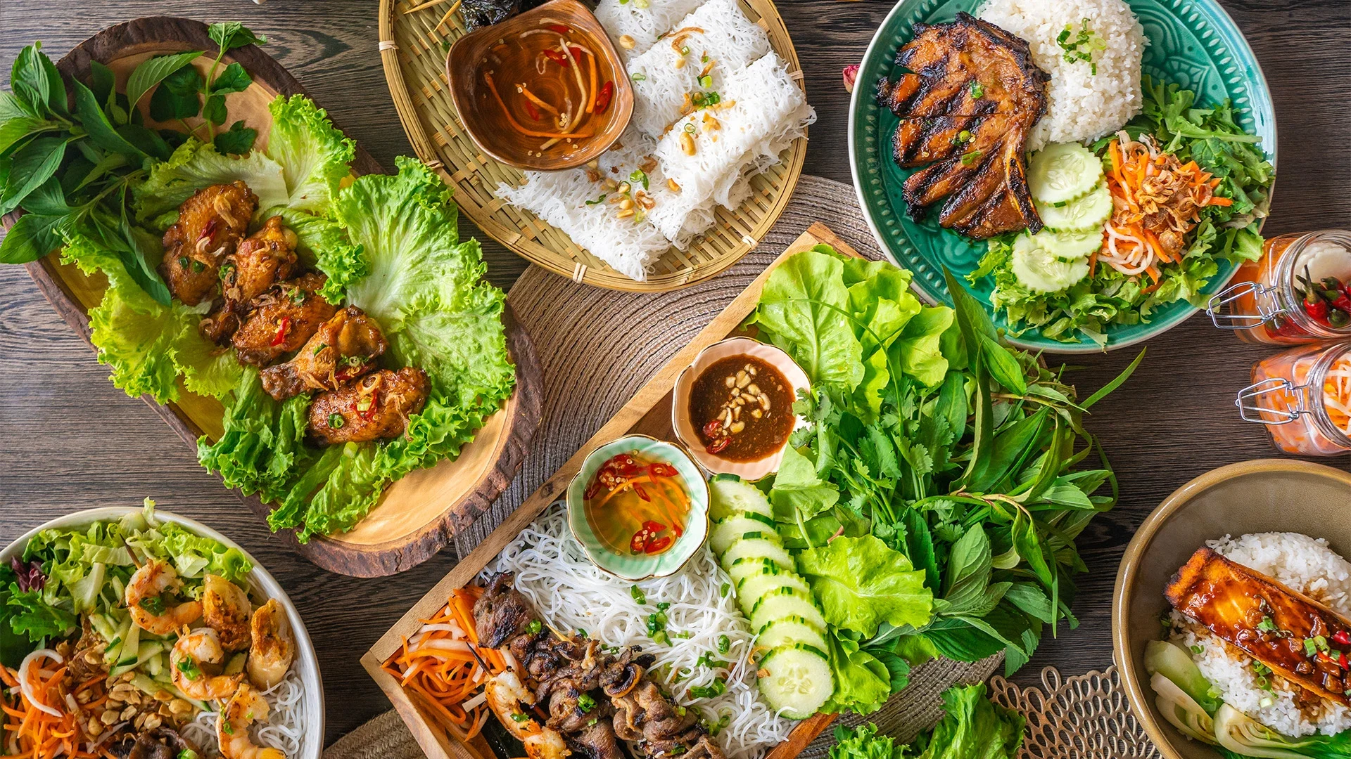 Assorted Vietnamese dishes on a wooden table, including grilled pork, rice rolls, rice with grilled fish, noodles, and fresh vegetables.