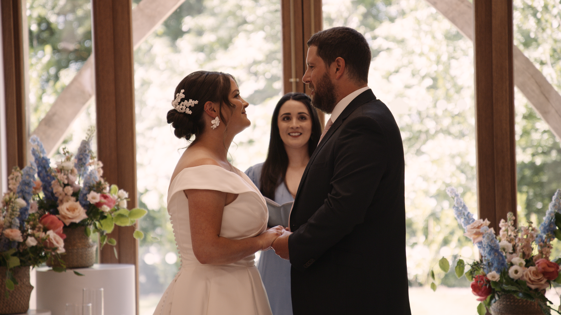 Couple at the alter with celebrant behind them. The bride is wearing a white dress and her hair is in an up-do. The groom is in a black suit.